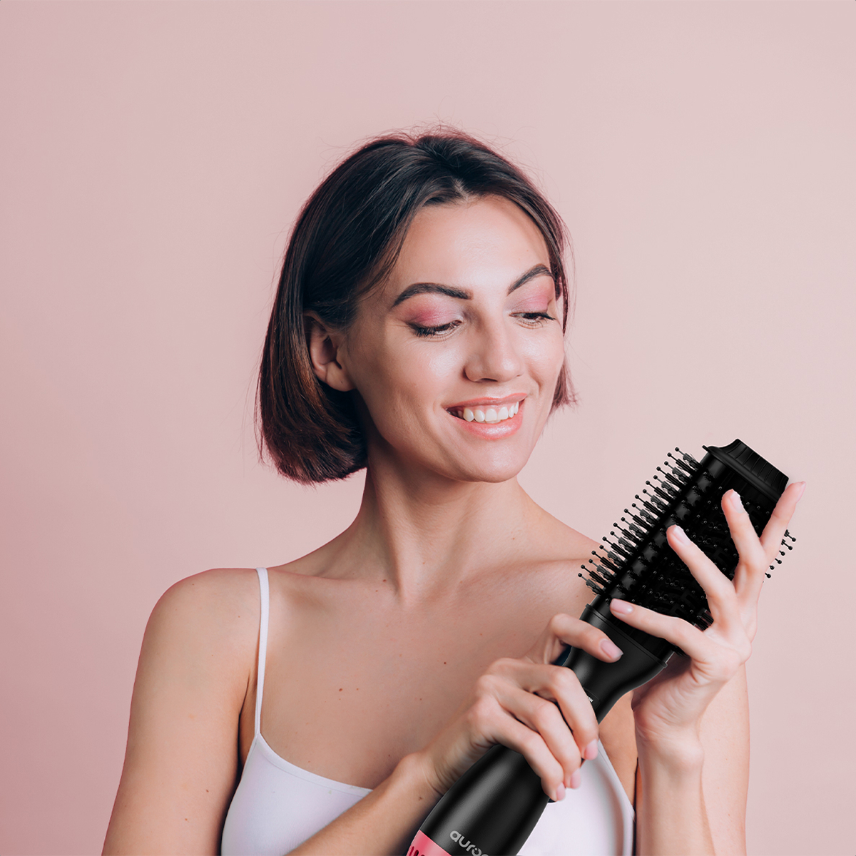 Woman holding a hair styler, smiling. Pink background.