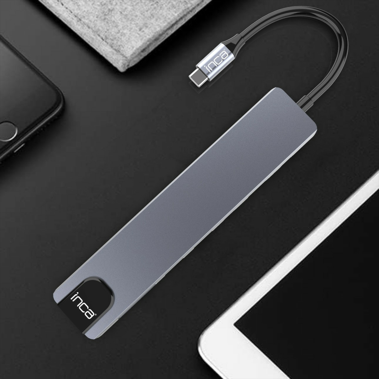 Un hub USB-C Inca grigio con cavo collegato, appoggiato su una scrivania accanto a un telefono e un tablet.