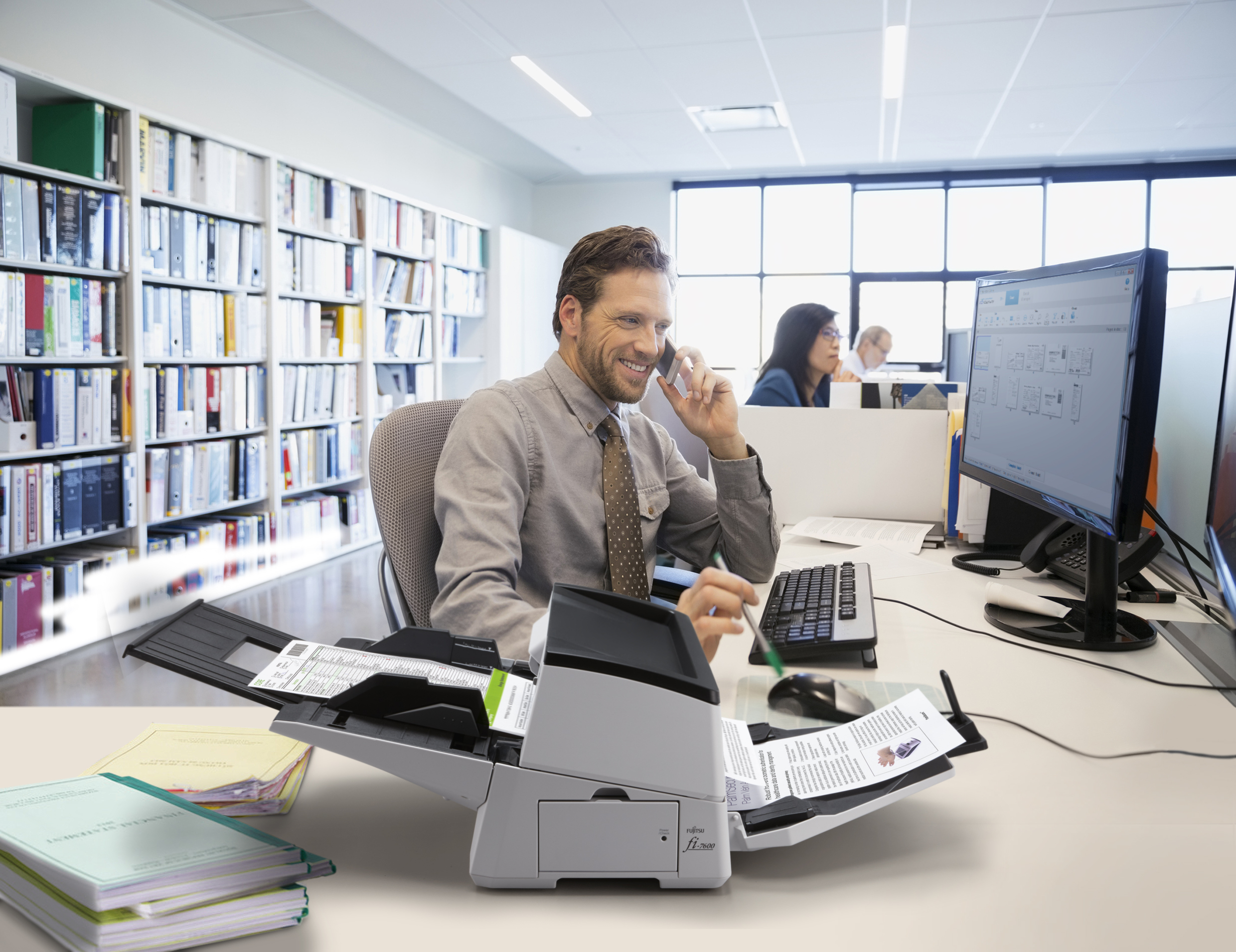 Mężczyzna w biurze używający skanera, rozmawiający przez telefon. Biurko z papierami i komputerem.