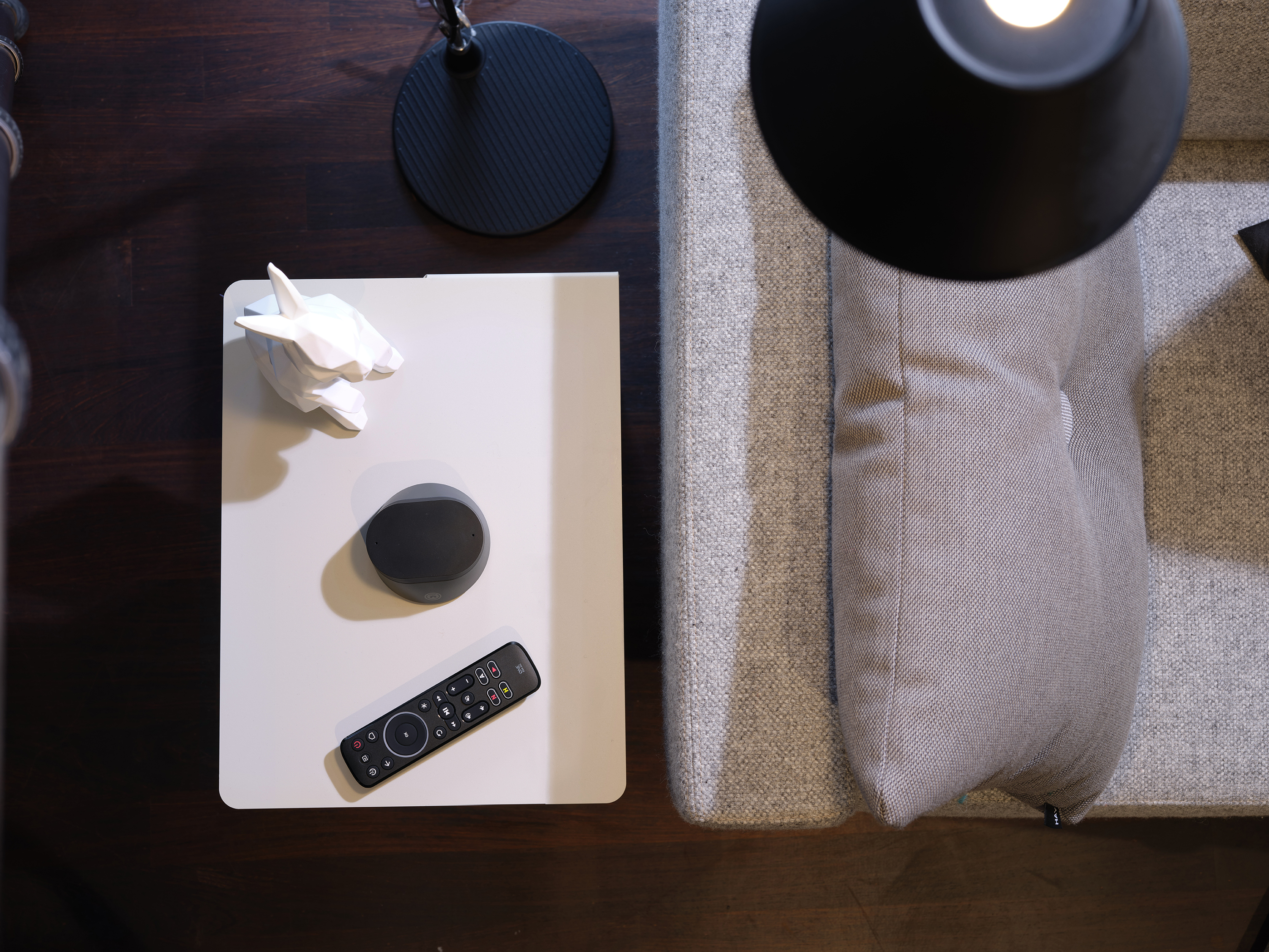 Widok z góry na stół z pilotem, czarną lampą i białą rzeźbą. Po prawej kanapa z poduszką.