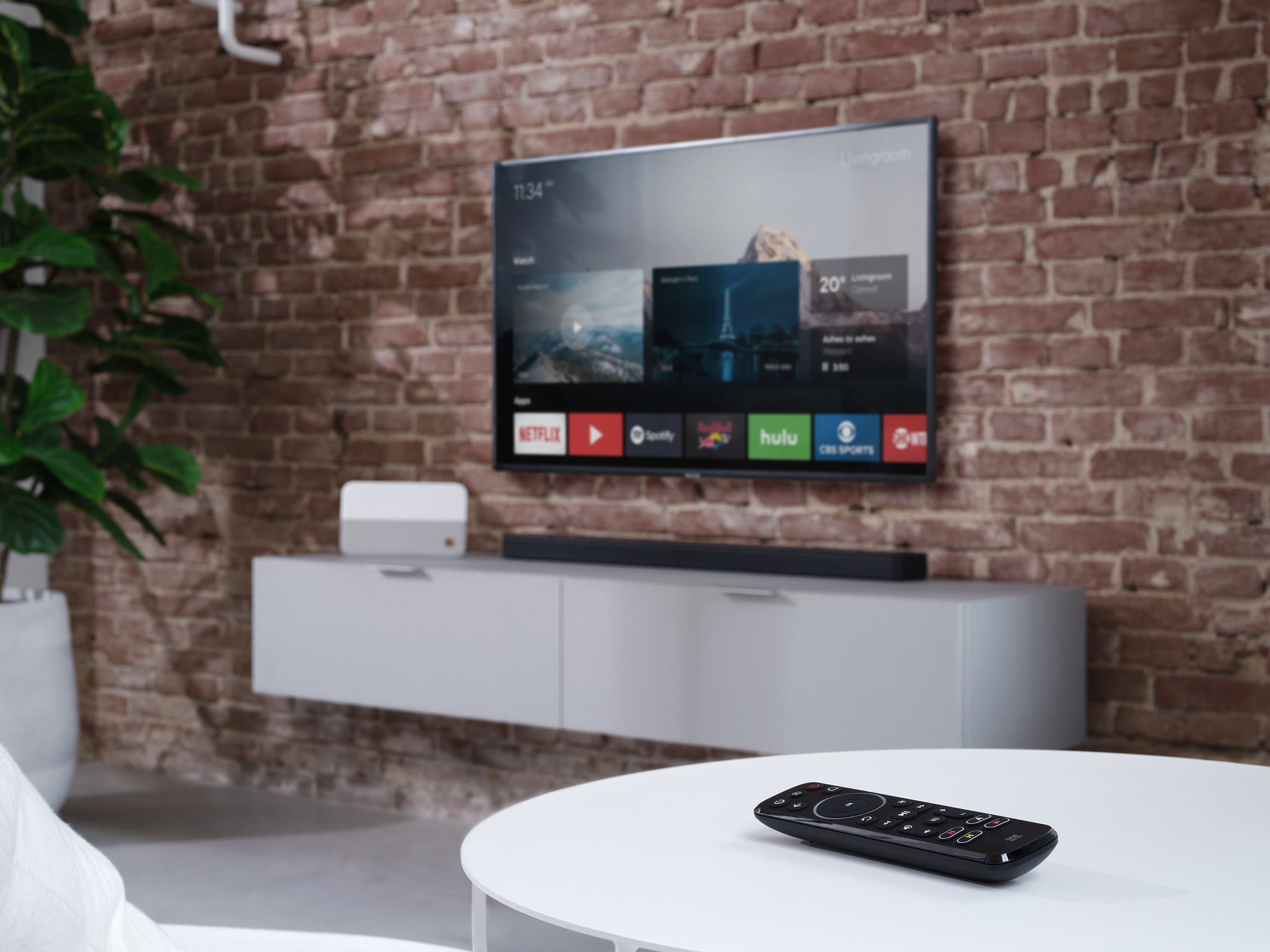 Telewizor na ścianie, z soundbarem i białym stołem z pilotem. Roślina po lewej stronie.