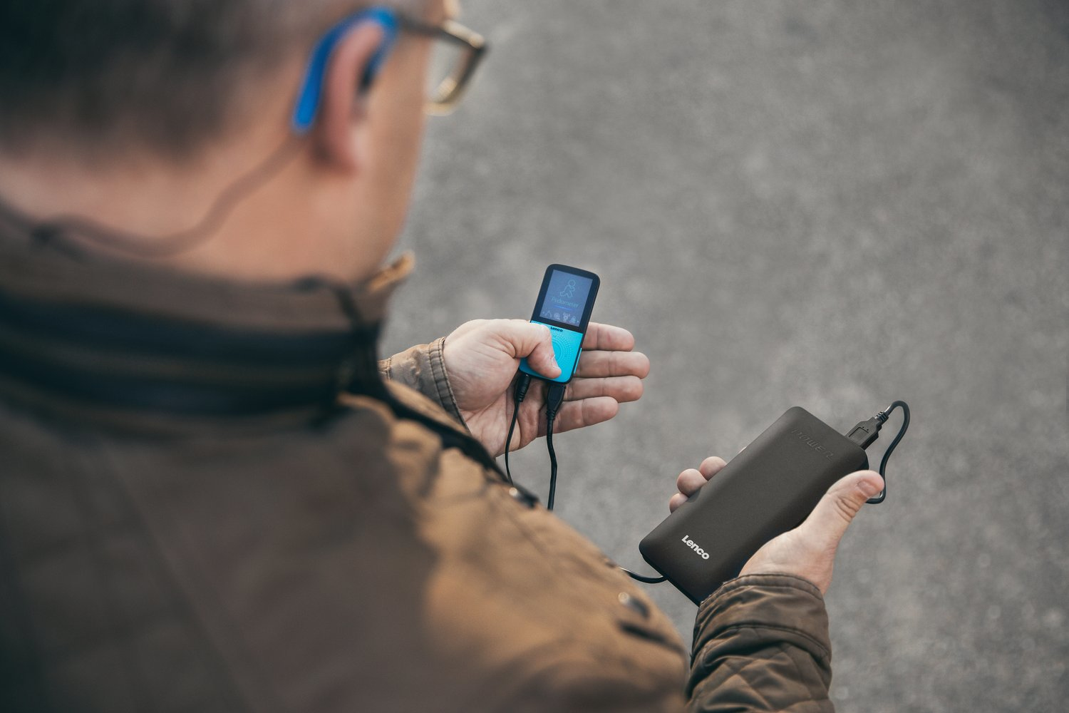 Mann mit Brille, hält einen türkisfarbenen MP3-Player und eine schwarze Powerbank. Verschwommener Hintergrund.