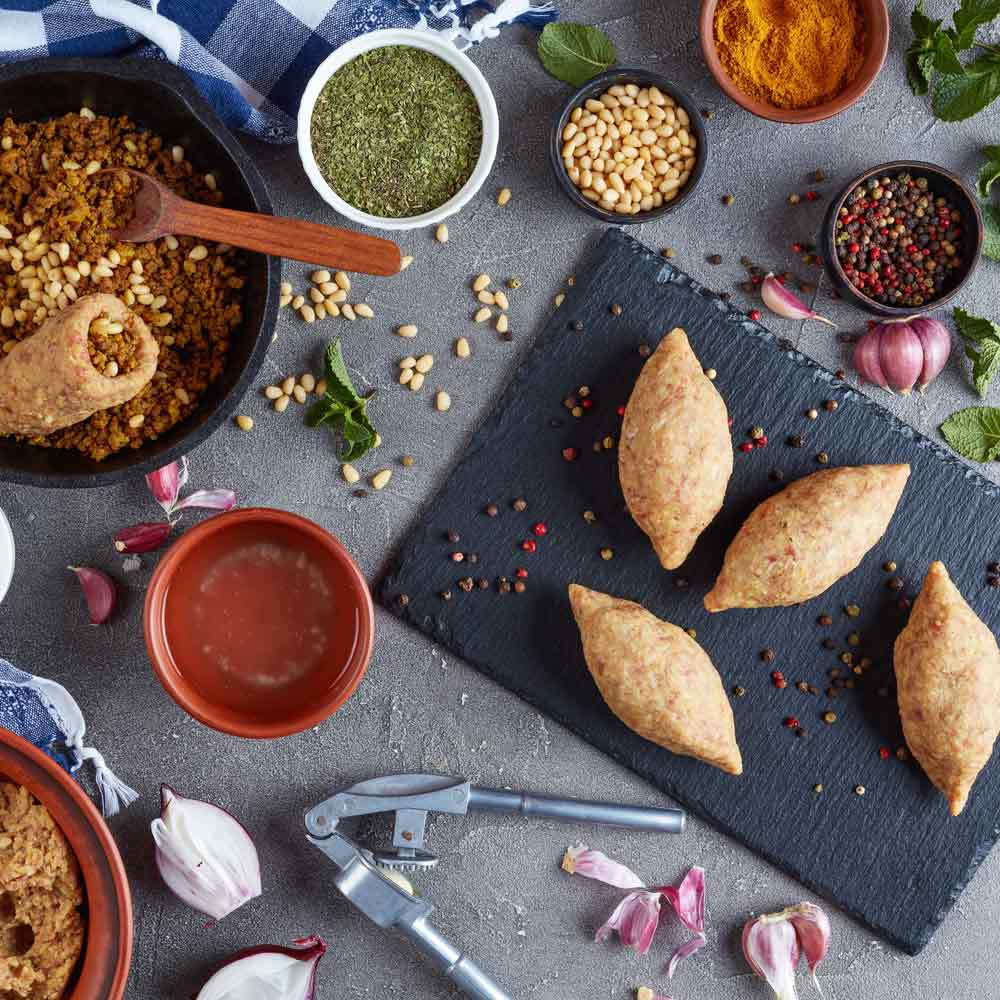 Stół z różnymi potrawami: kibbeh, orzeszki piniowe, przyprawy i warzywa, ułożone na szarej powierzchni.