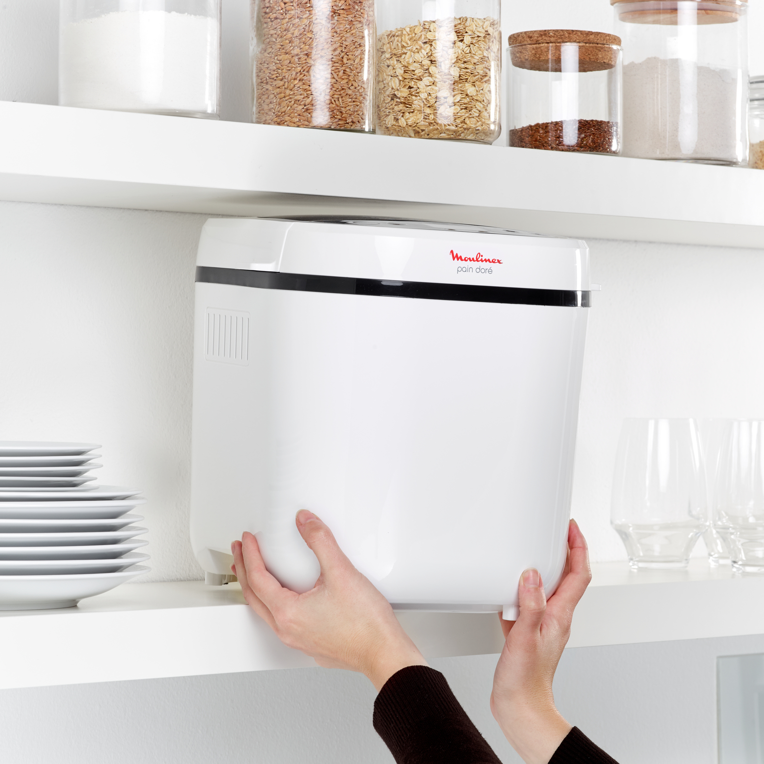 Biała maszyna do chleba Moulinex na półce, trzymana przez dwie ręce, w pobliżu szklanych słoików i talerzy.