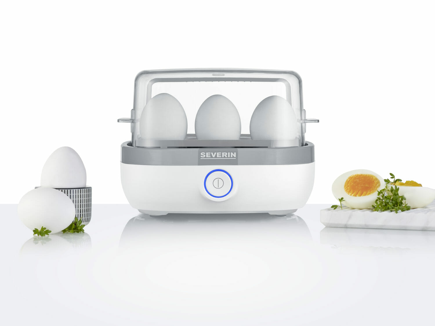Biało-szara parowar do jajek z trzema jajkami w środku, otoczona jajkami i dodatkami.