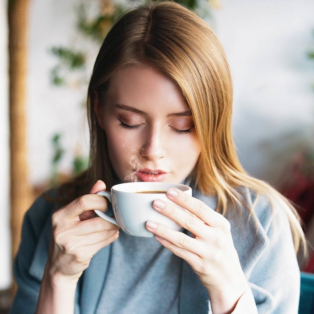 Una donna tiene una tazza di caffè, sale vapore. Ha i capelli biondi e indossa un top grigio.