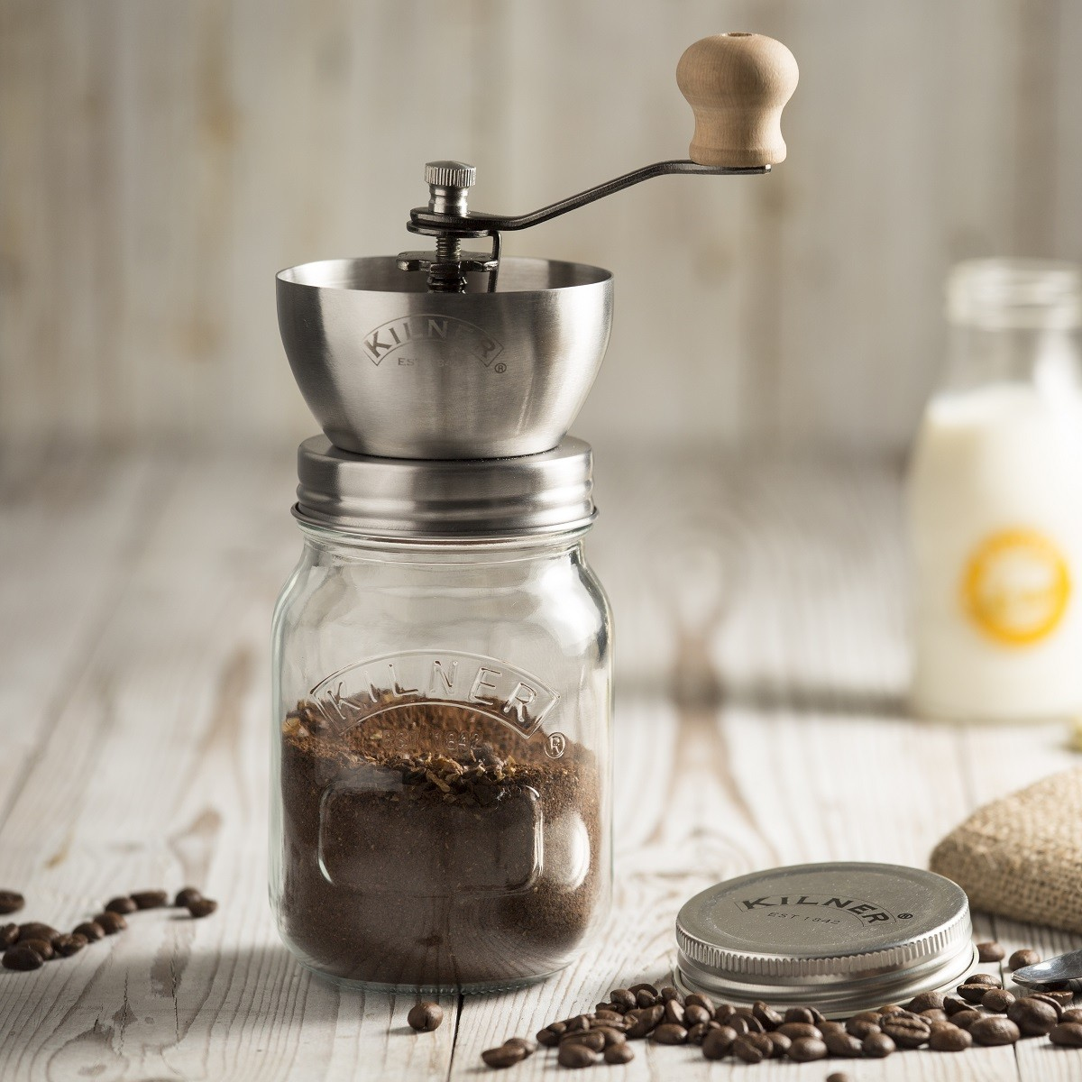 Eine manuelle Kaffeemühle auf einem Glas mit gemahlenem Kaffee, Kaffeebohnen und einer Milchflasche.