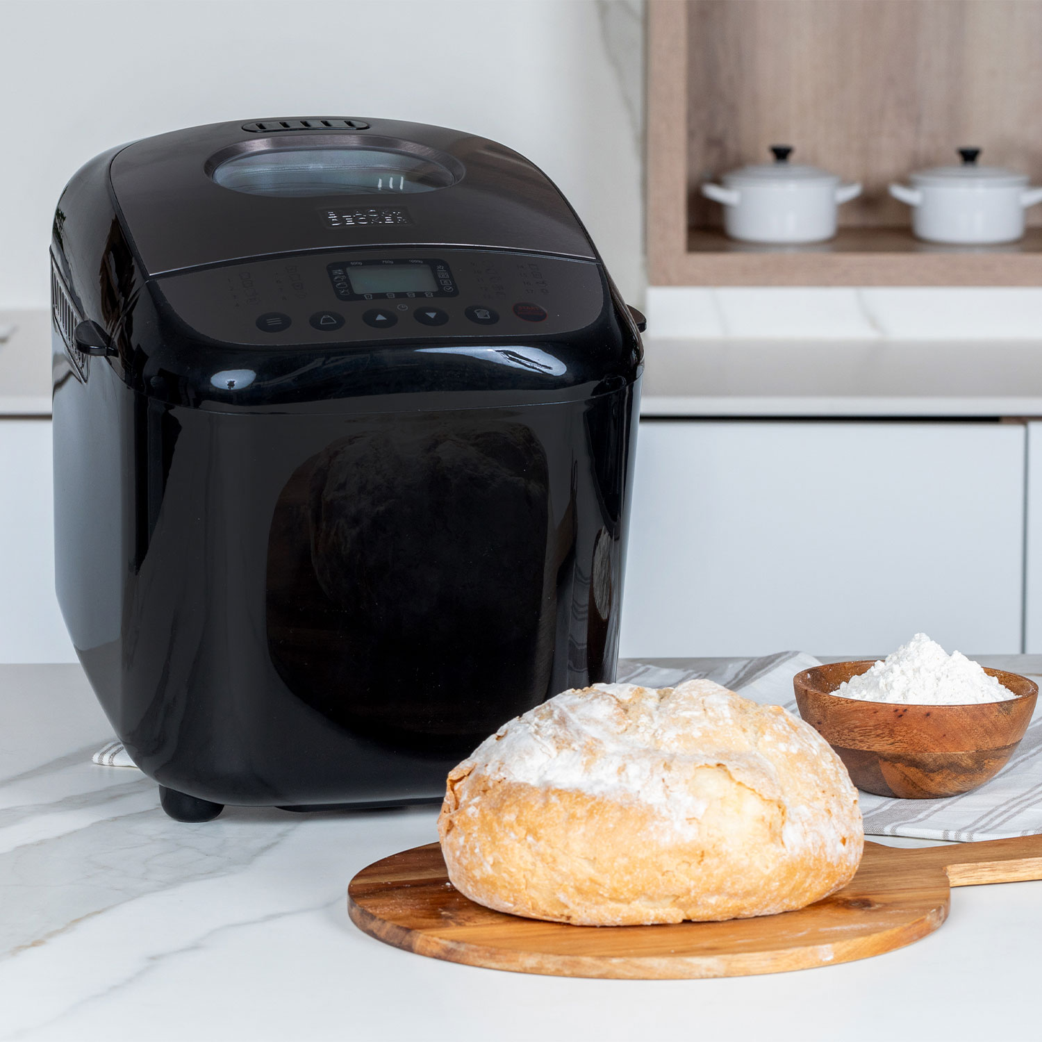 Een zwarte broodbakmachine met een brood en ingrediënten op een tafel.