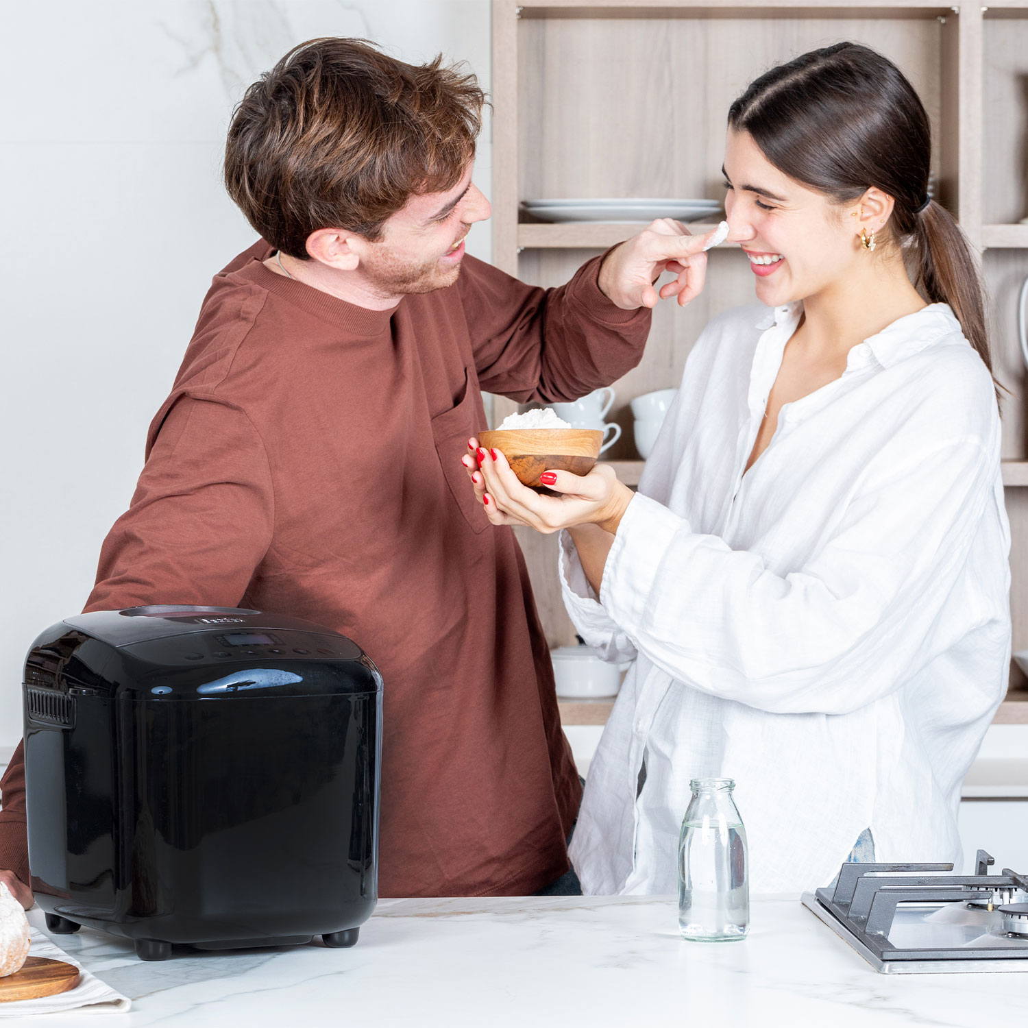 Een stel in een keuken met een broodbakmachine. Man doet bloem op de neus van de vrouw.