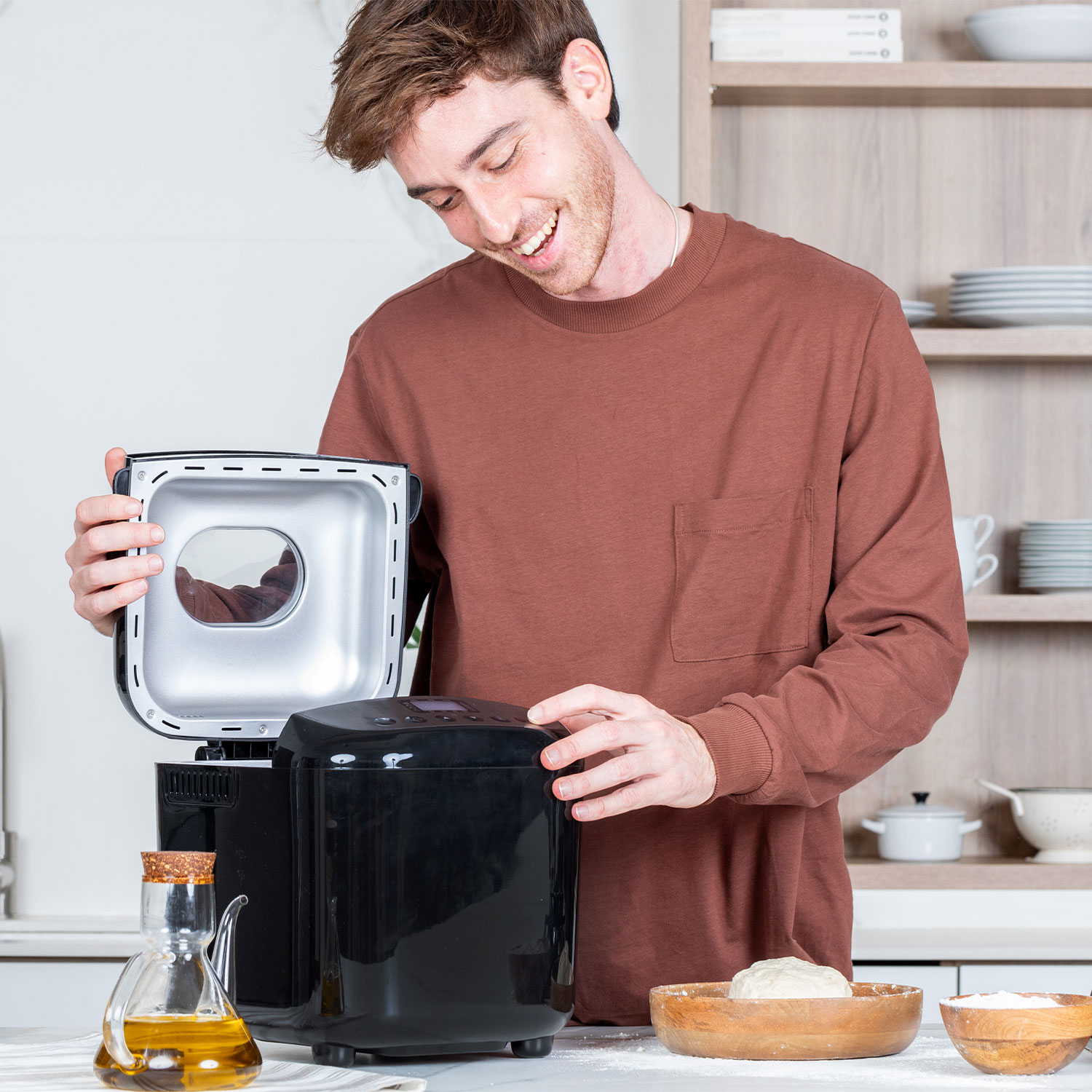 Man opent de bovenkant van een zwarte broodbakmachine. Ingrediënten in kommen op het aanrecht.