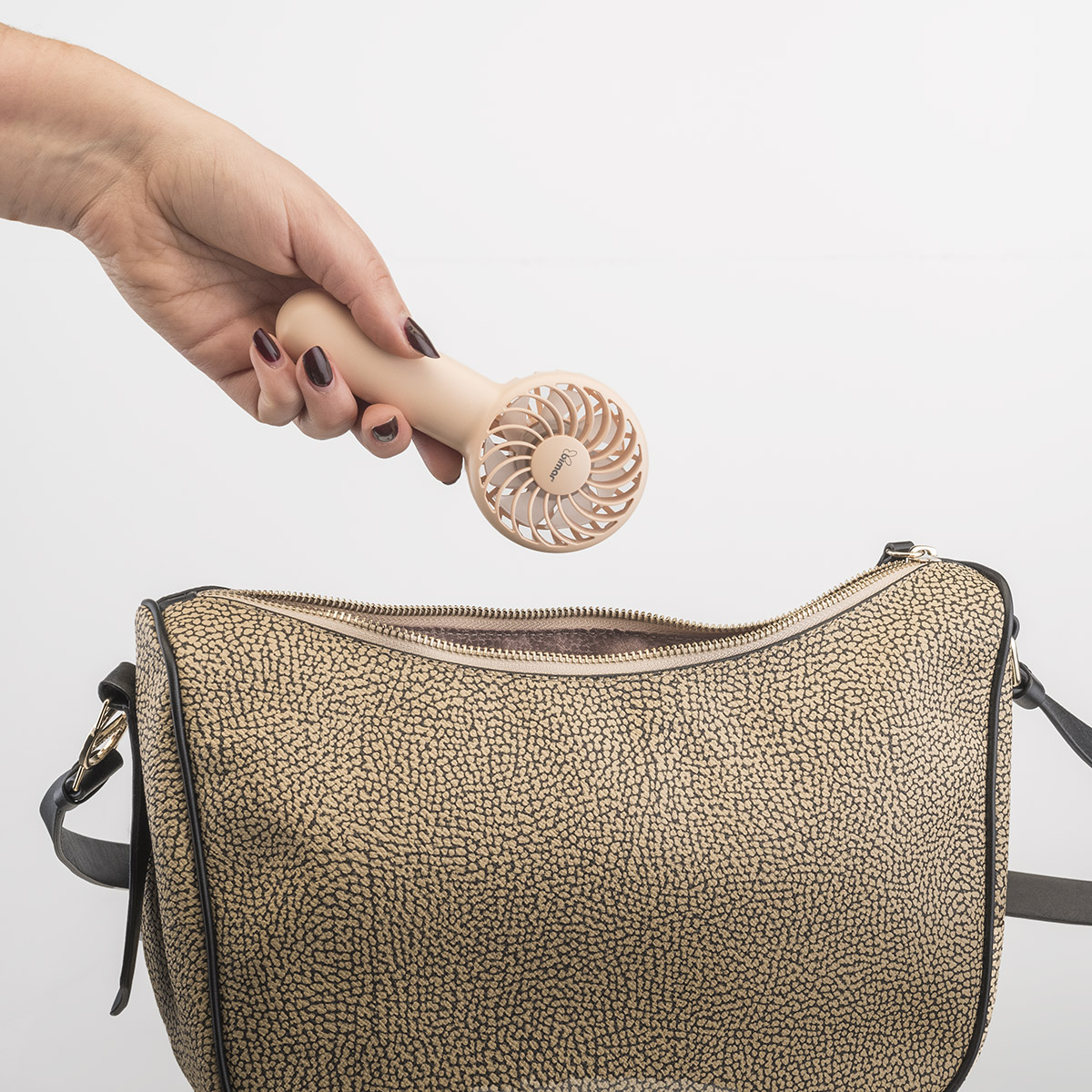 Mano che tiene un piccolo ventilatore portatile beige, sopra una borsa beige e nera.