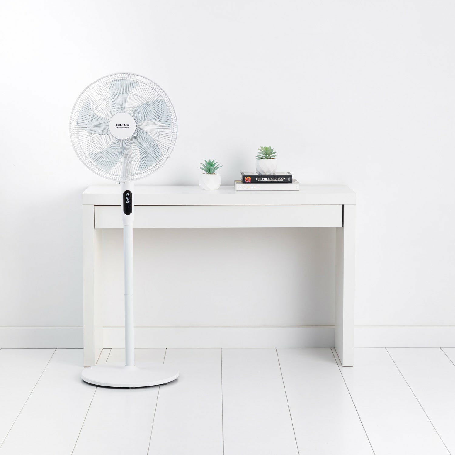 Ventilador blanco sobre un soporte y mesa blancos. Dos pequeñas plantas y un libro en la mesa.