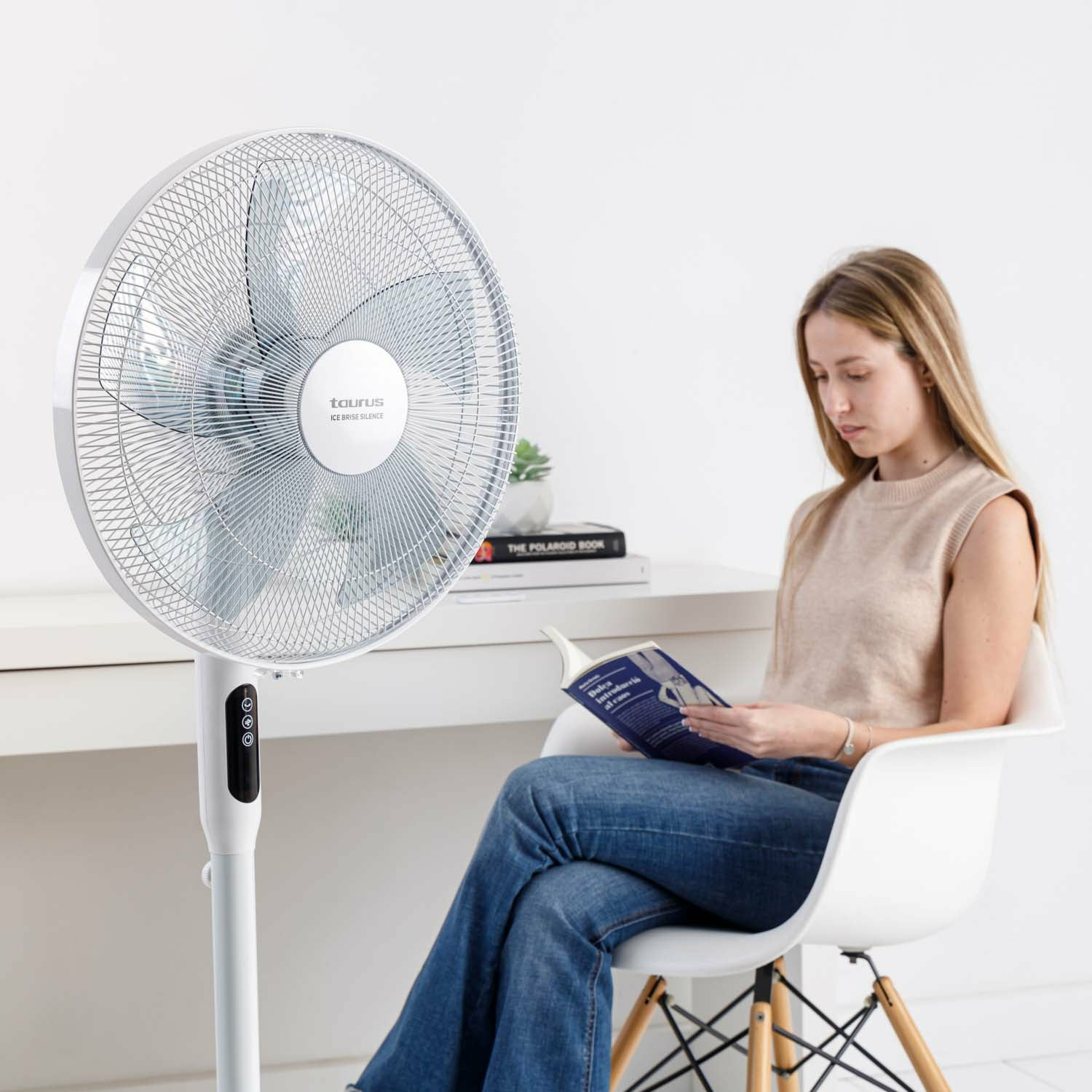Ventilador de pie blanco y plateado, con una mujer leyendo un libro, fondo blanco.