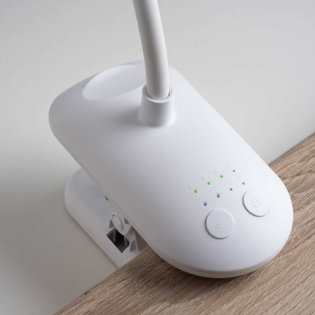Una lampada da scrivania bianca con collo flessibile. Ha una base a clip e comandi touch. Su una superficie in legno.