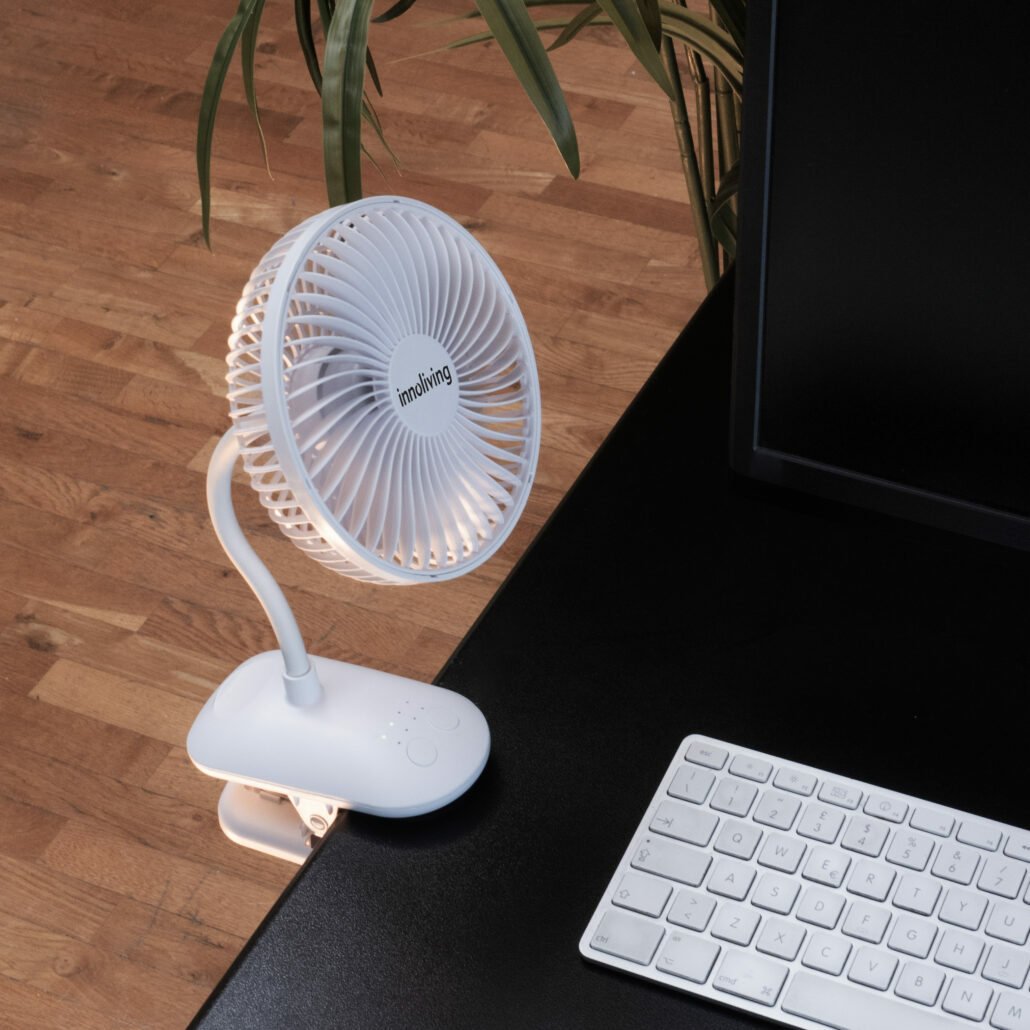 Ventilatore bianco agganciato a una scrivania nera, accanto a una tastiera. Pavimento in legno.