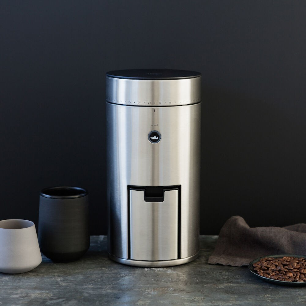 Kaffeemühle aus Edelstahl, mit Kaffeetasse und Bohnen daneben.
