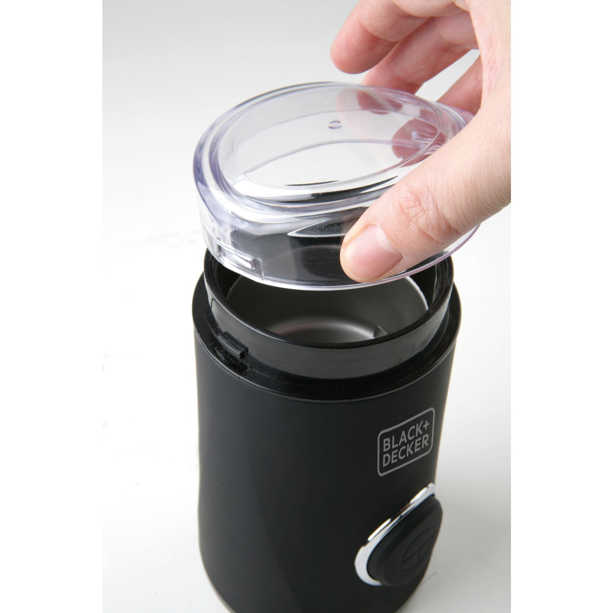 Czarny młynek do kawy z przezroczystą pokrywą. Ręka trzyma pokrywę. Białe tło.