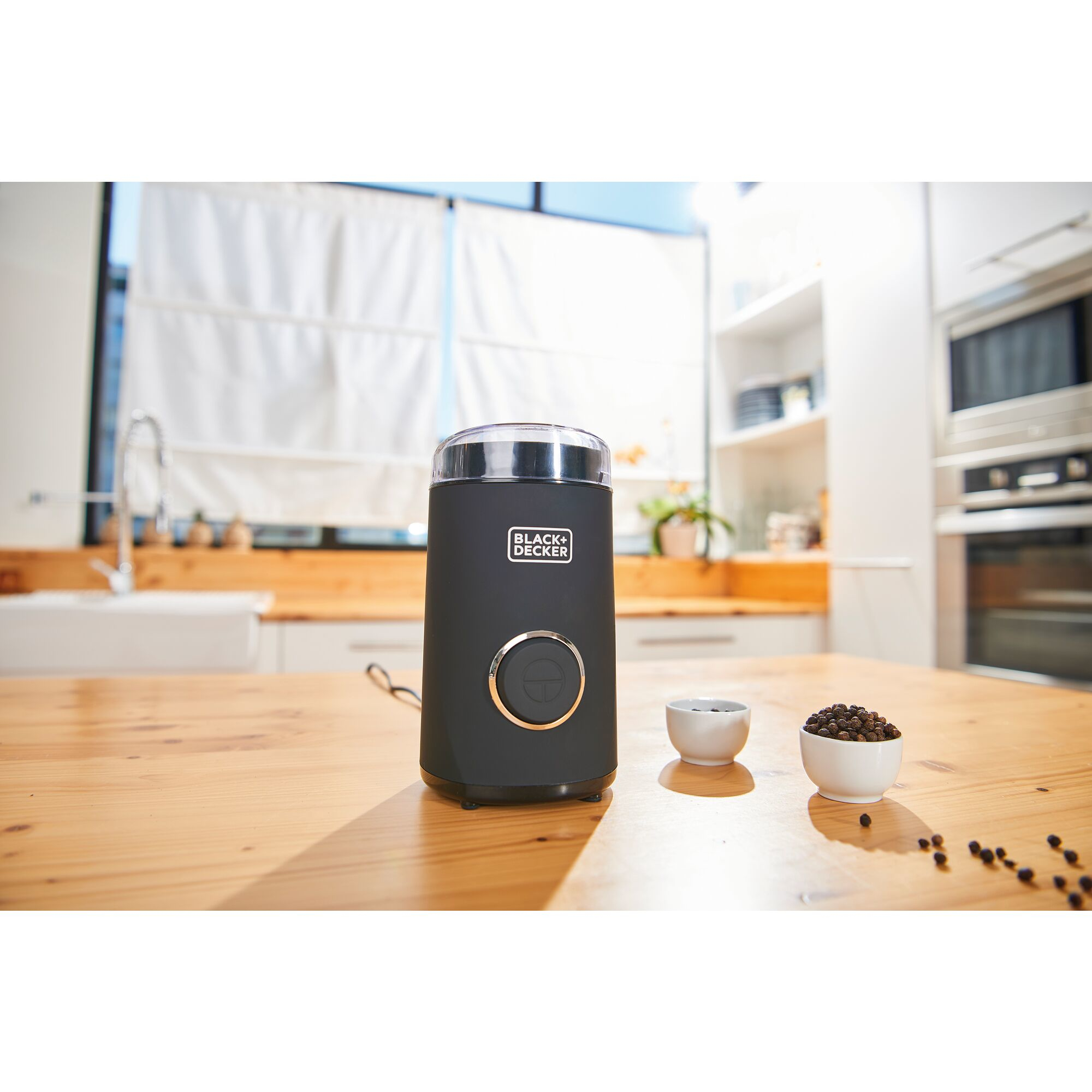 Czarny młynek do kawy ze złotym guzikiem, obok małych miseczek z ziarnami kawy, na drewnianym stole.