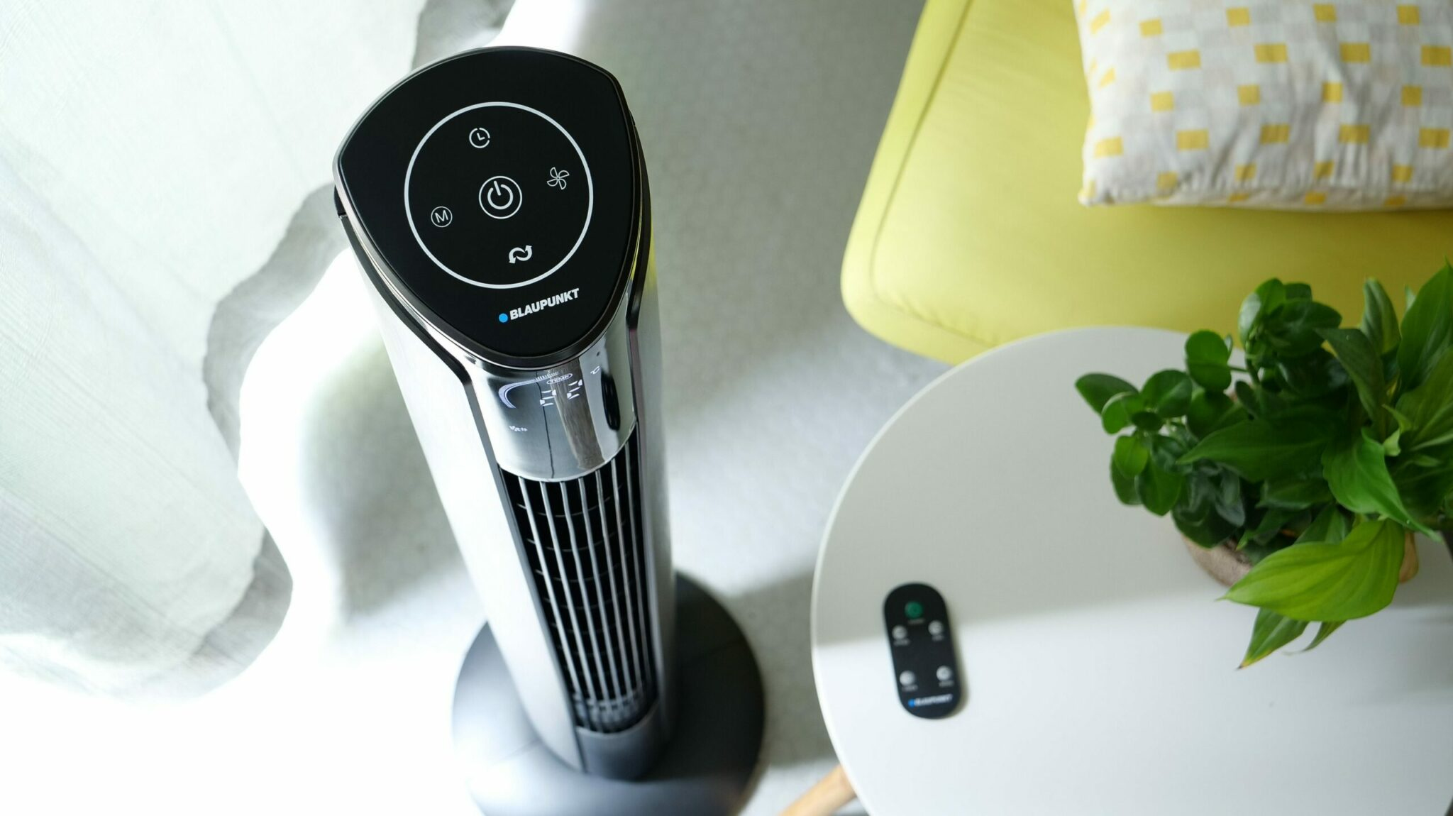 Ventilador alto con panel de control negro, mesa con planta y mando, silla amarilla.