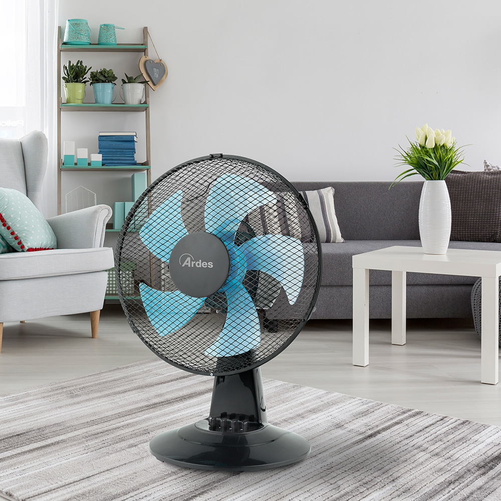Wentylator stołowy na dywanie. Niebieskie łopatki, czarna siatka. Szara kanapa i biały stół w tle.