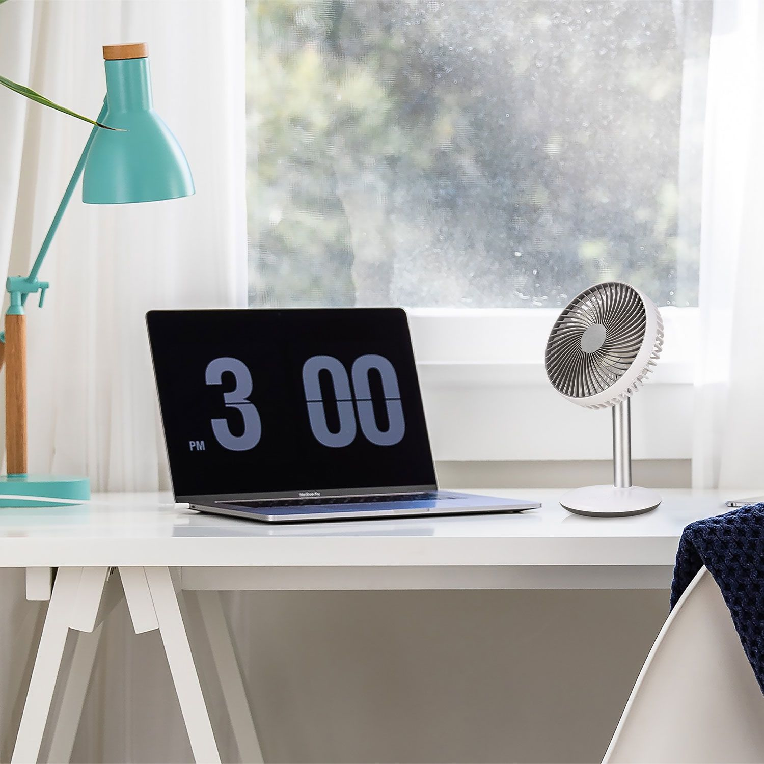 Schreibtisch mit Laptop (15:00 Uhr), Ventilator, Lampe und Fenster im Hintergrund.
