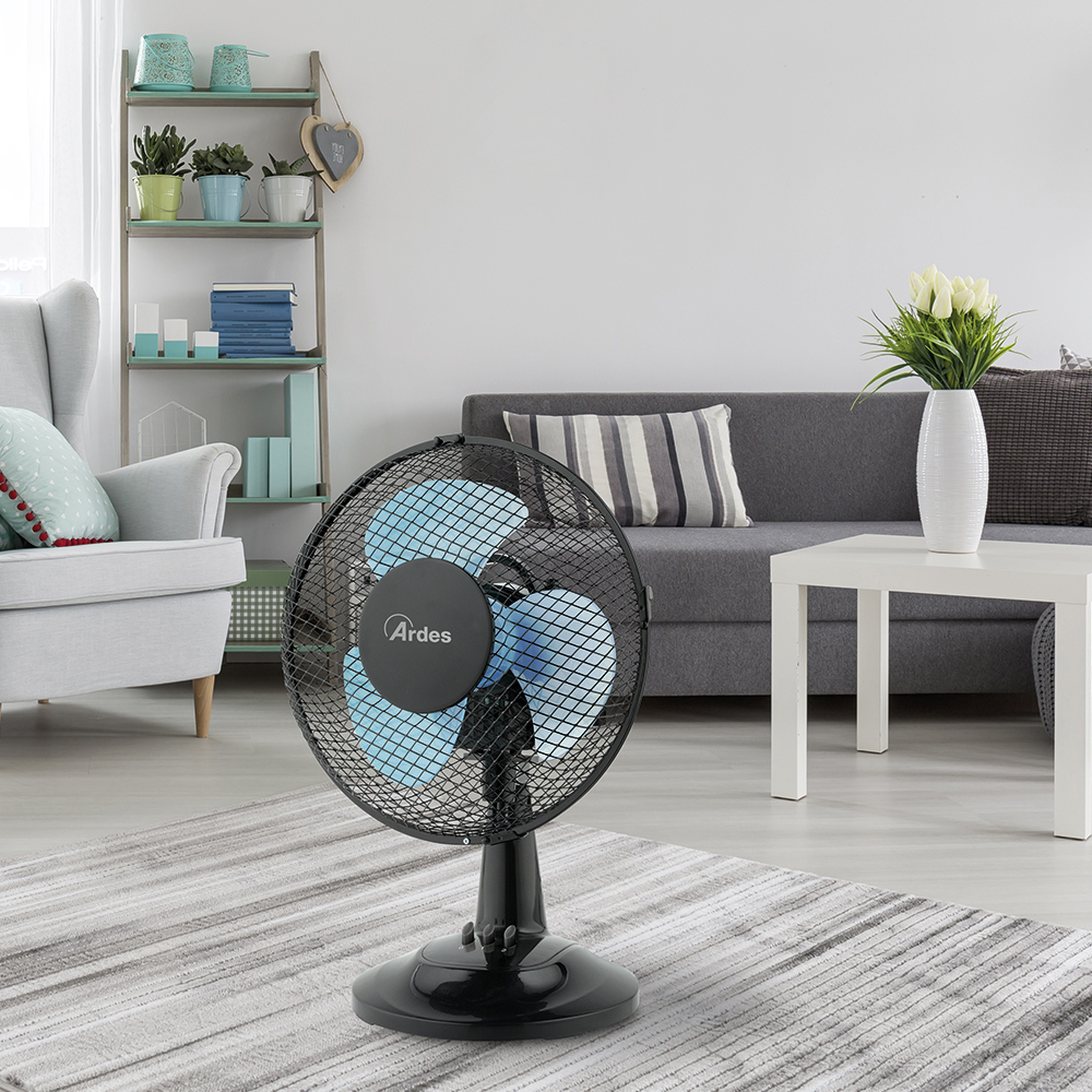 Een zwarte ventilator op een gestreept tapijt, in een woonkamer met een bank en een plank.