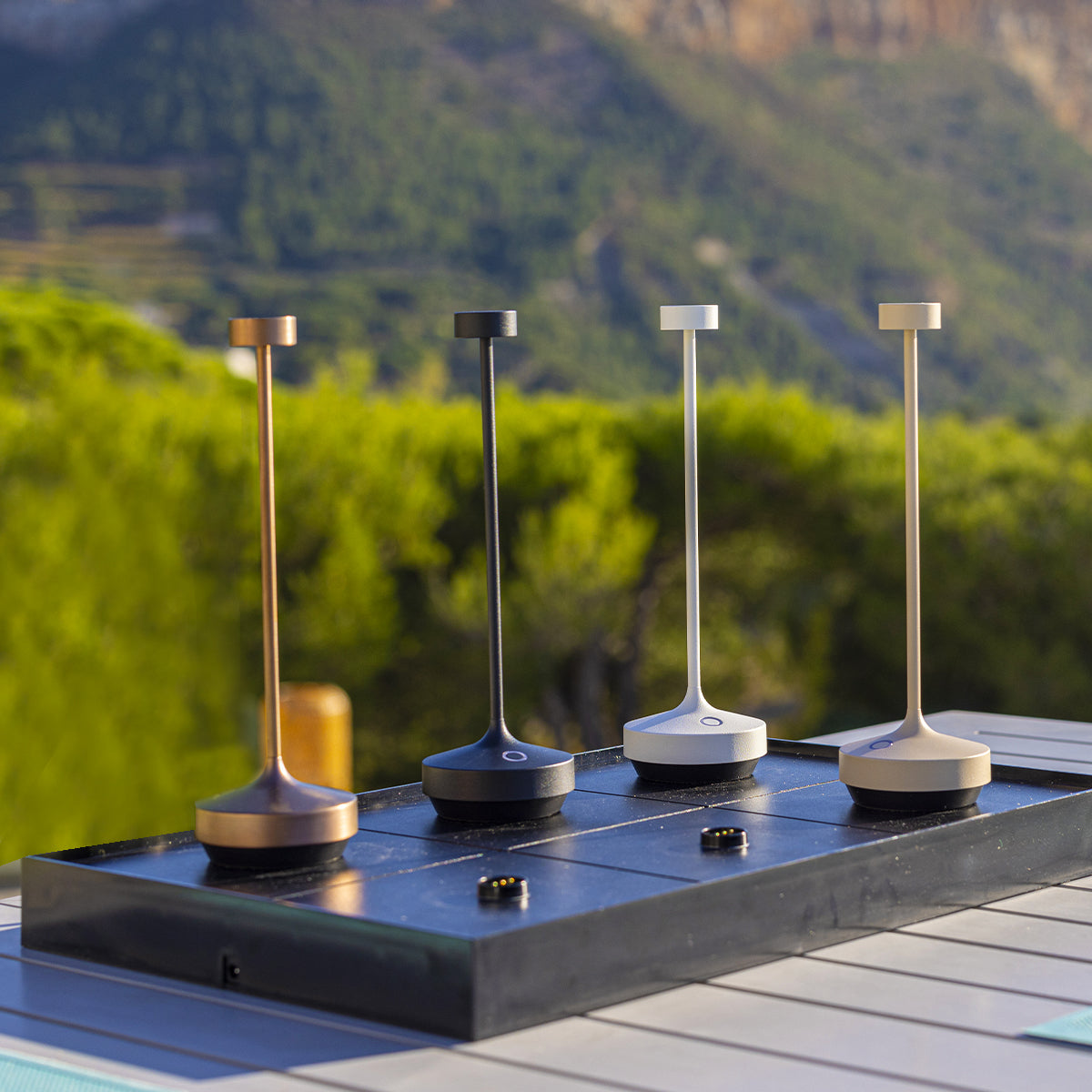 Vier minimalistische Tischlampen in verschiedenen Farben auf schwarzer Oberfläche, mit Bergkulisse.