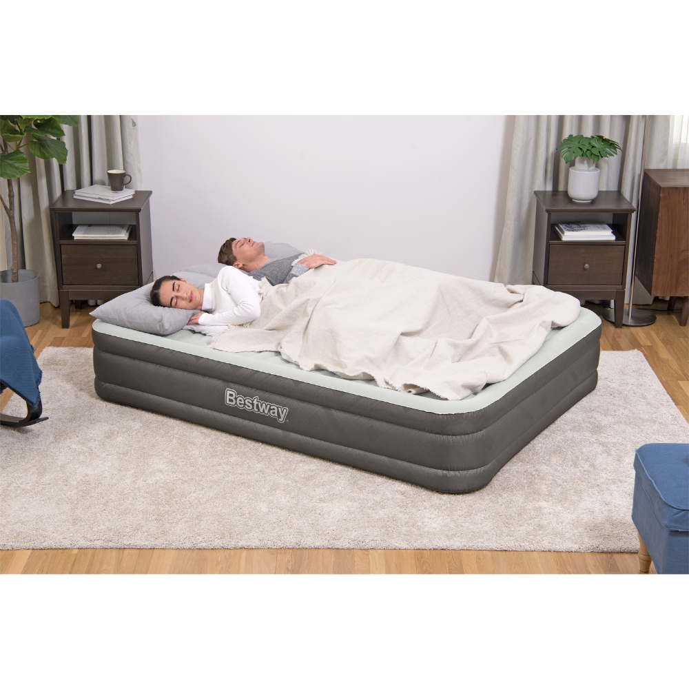 Deux personnes sont allongées sur un matelas pneumatique gris dans une pièce avec des tables de chevet. Il y a un tapis.