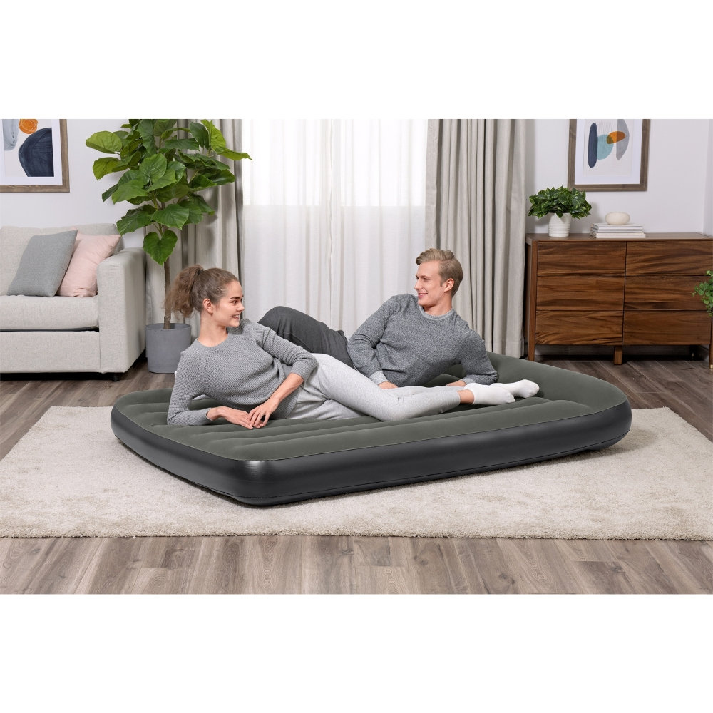 Un couple est allongé sur un matelas gonflable gris dans une pièce. Arrière-plan : tapis, meubles et rideaux.