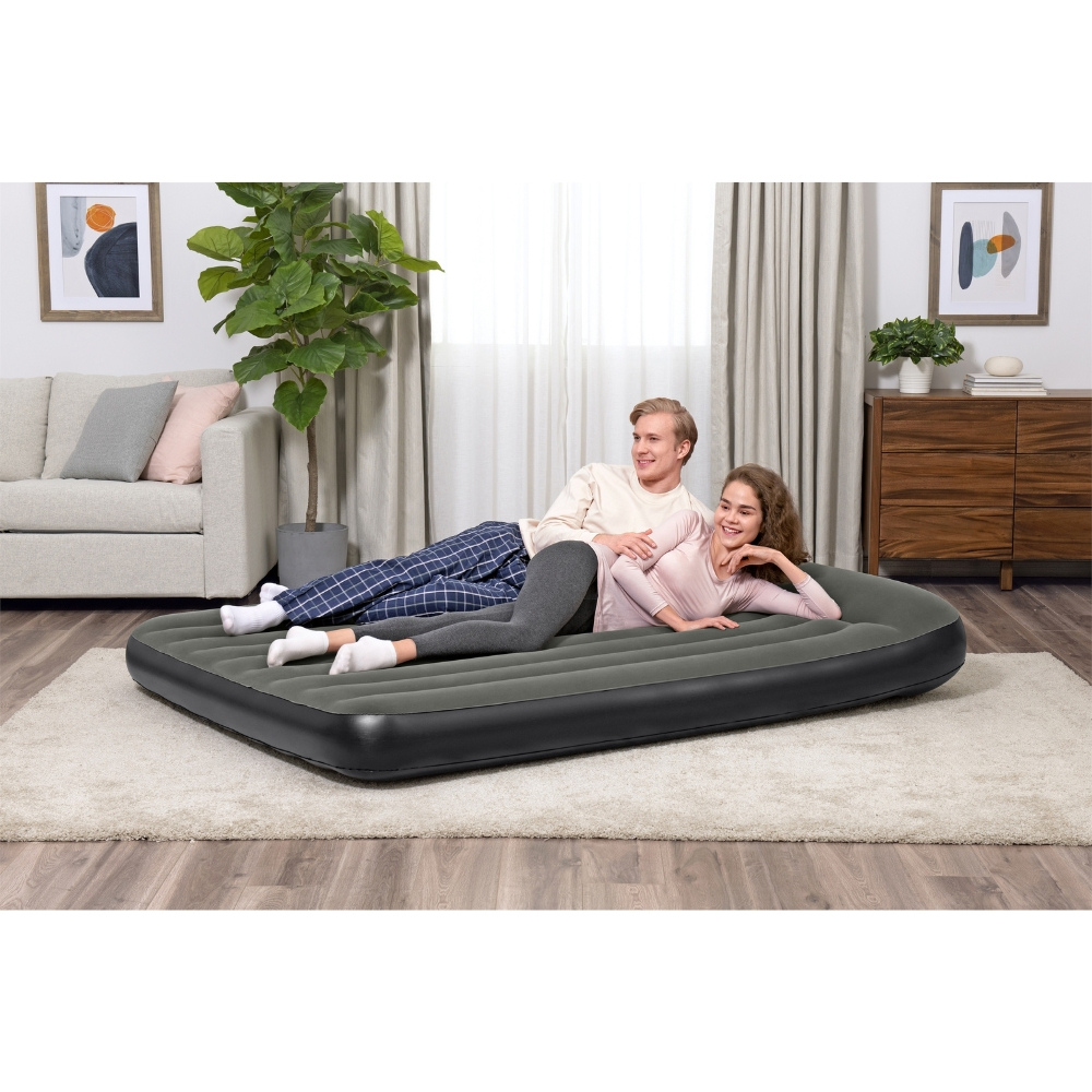 Un couple est allongé sur un matelas gonflable gris dans une pièce. Ils sourient. Arrière-plan : meubles, rideaux.