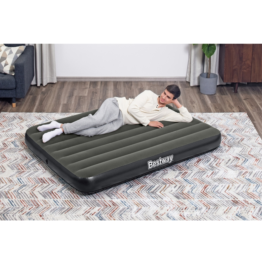 Un homme est allongé sur un matelas gonflable gris foncé sur un tapis à motifs. Un canapé est en arrière-plan.
