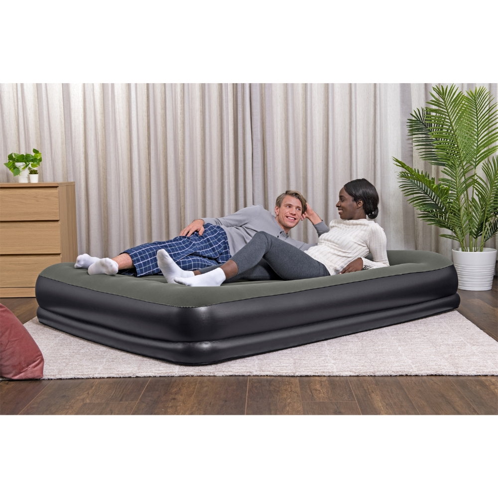 Un couple est allongé sur un matelas gonflable gris foncé sur un tapis. Une plante et une commode sont en arrière-plan.