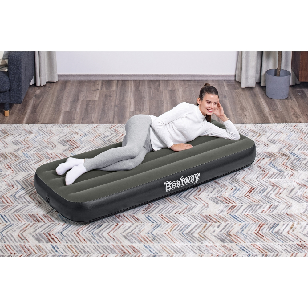 Une femme est allongée sur un matelas gonflable gris, souriante. Le logo Bestway est visible. Arrière-plan : un tapis.