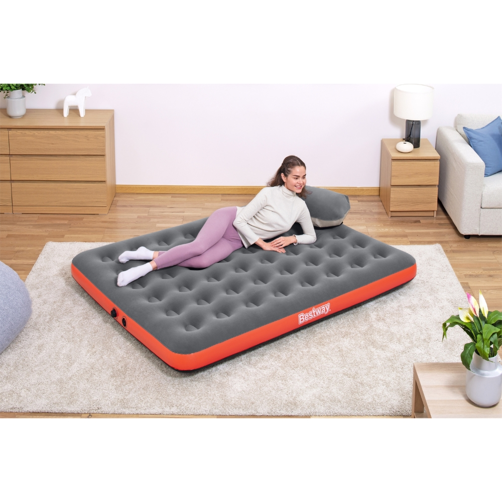 Een vrouw ligt op een grijze en oranje opblaasbare matras en glimlacht. Er is een Bestway-logo en een kussen.