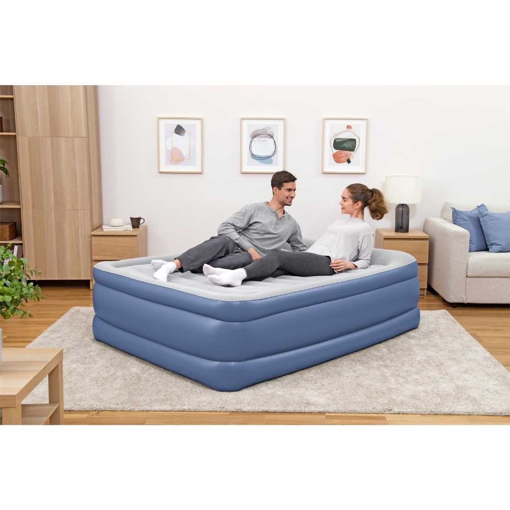 Couple sur matelas gonflable bleu, homme et femme parlent, tapis beige, meubles en bois.