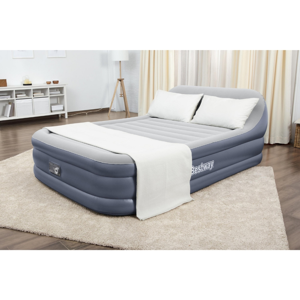 Een opblaasbare matras met kussens en een deken staat in een kamer. Er is een tapijt, een lamp en andere meubels.