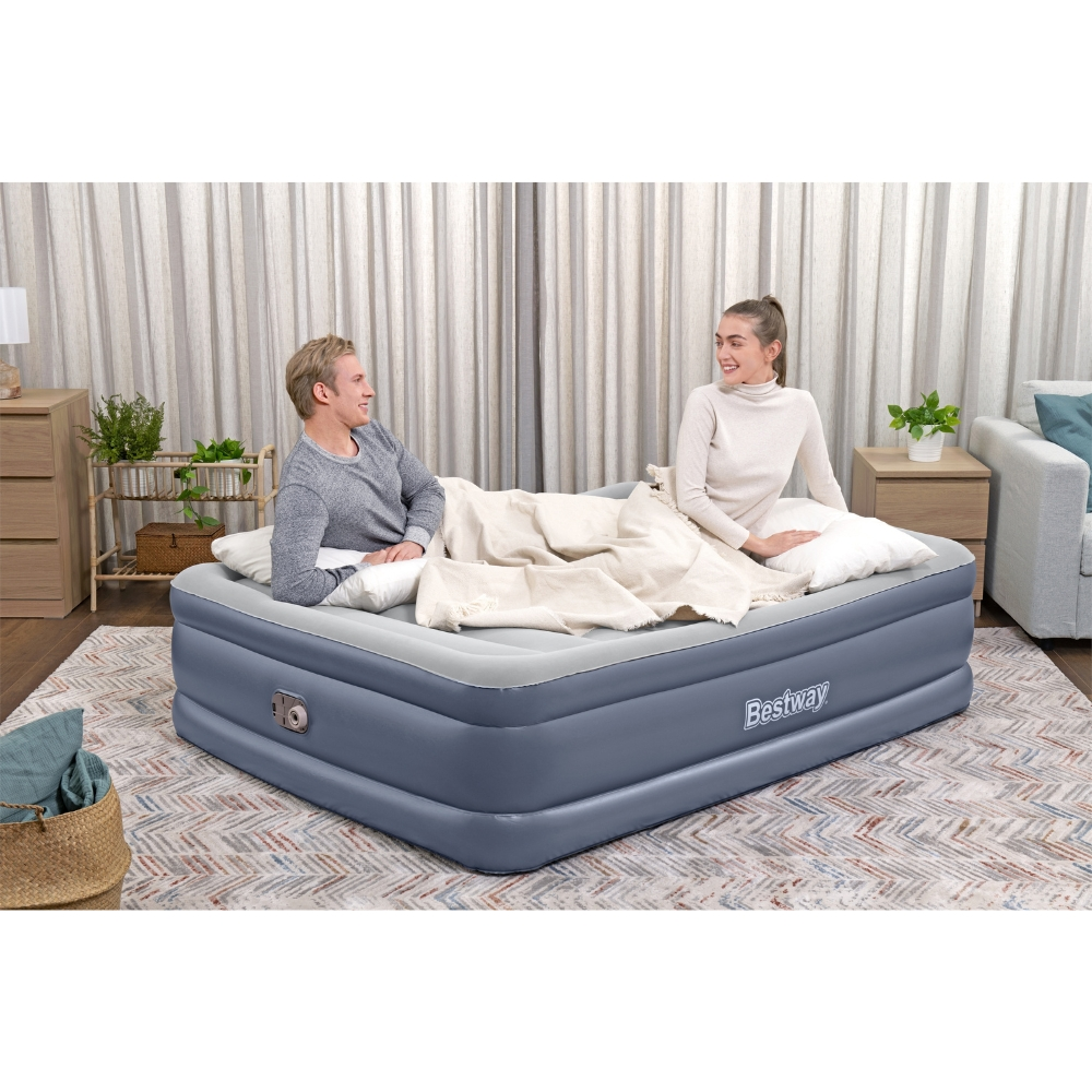 Een stel zit op een Bestway opblaasbare matras. Er liggen kussens en een deken op het bed.