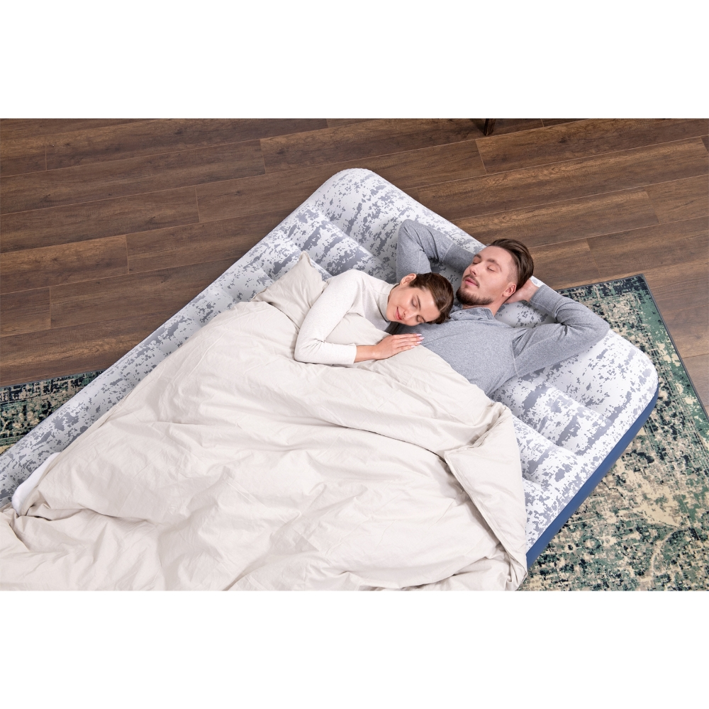 Couple dormant sur un matelas pneumatique à motifs, sous une couverture. Sol en bois sombre, tapis vert.