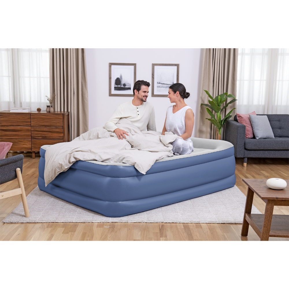 Een stel zit op een opblaasbare matras in een kamer. Er ligt een deken en kussens op het bed.