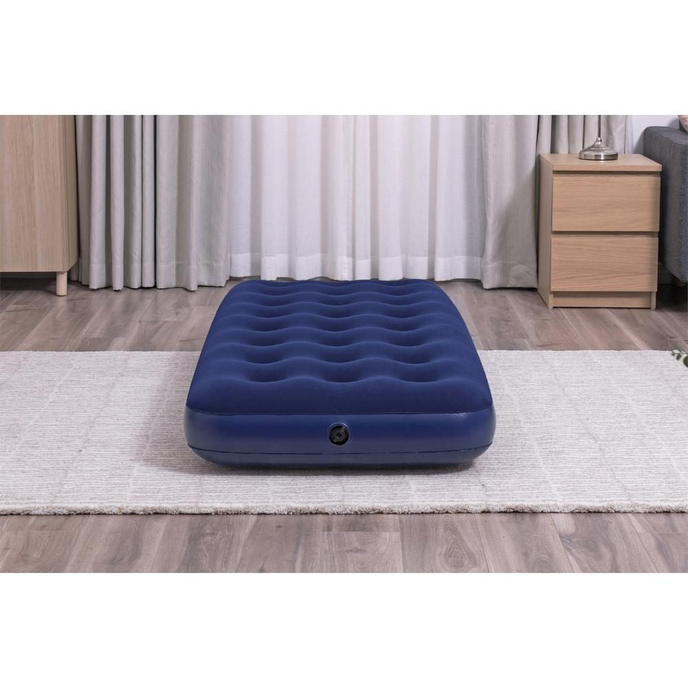 Een blauwe opblaasbare matras op een lichtgekleurd vloerkleed. Het is binnen, met een lamp en meubels op de achtergrond.