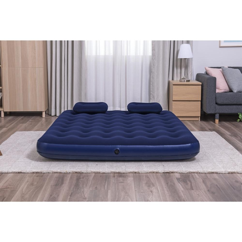 Matelas pneumatique bleu foncé avec deux oreillers sur un tapis. La pièce comprend des meubles en bois et une fenêtre.