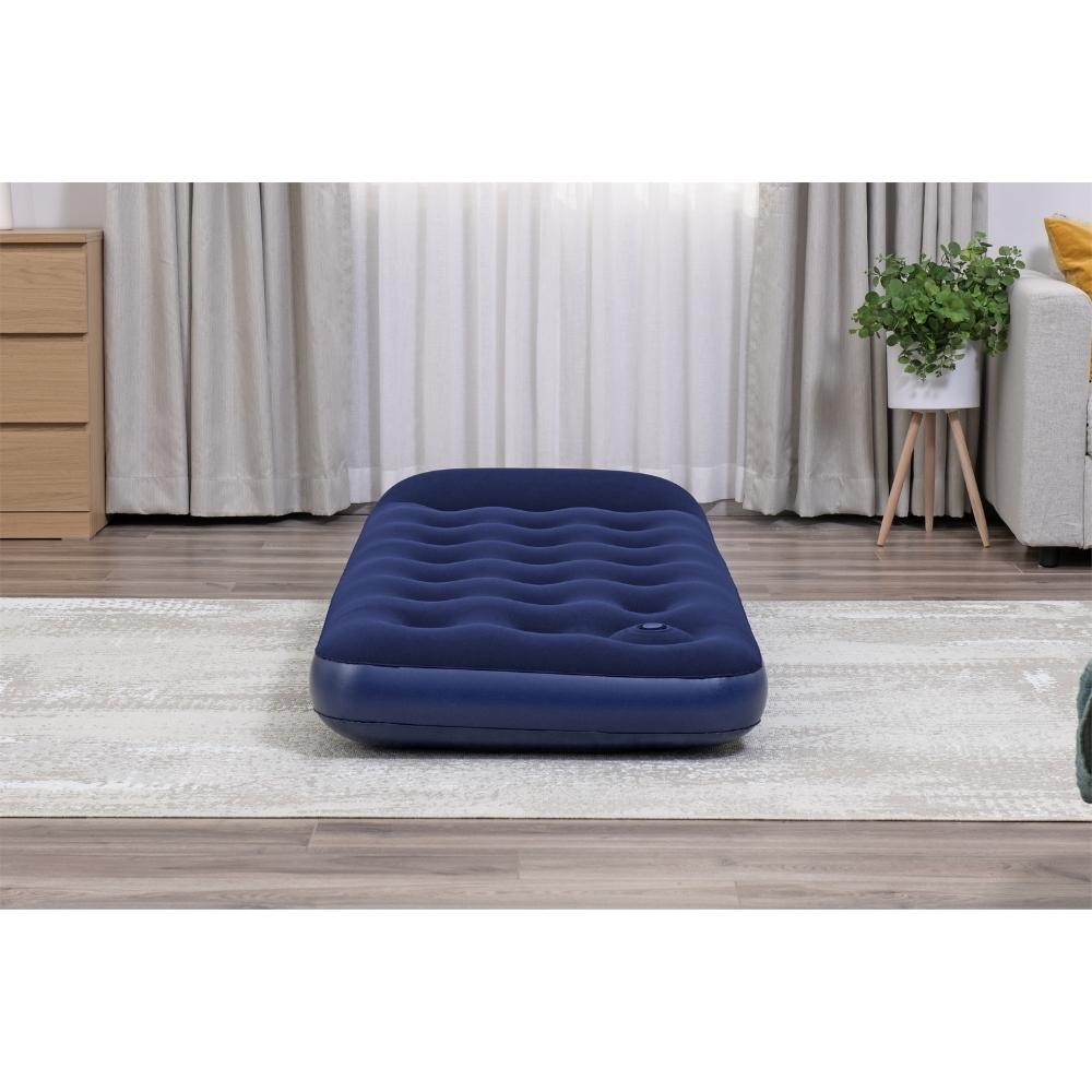 Blauw luchtbed in een kamer, op een grijs tapijt. Houten vloer, beige gordijnen en een klein kastje.