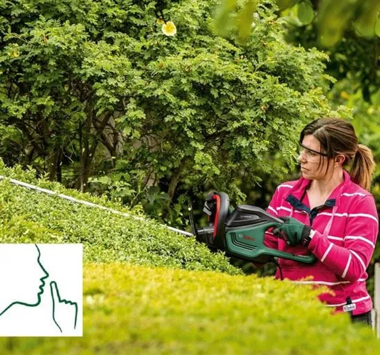 Frau schneidet Hecke mit grün-schwarzem Elektro-Heckenschneider. Grüne Pflanzen umgeben sie.