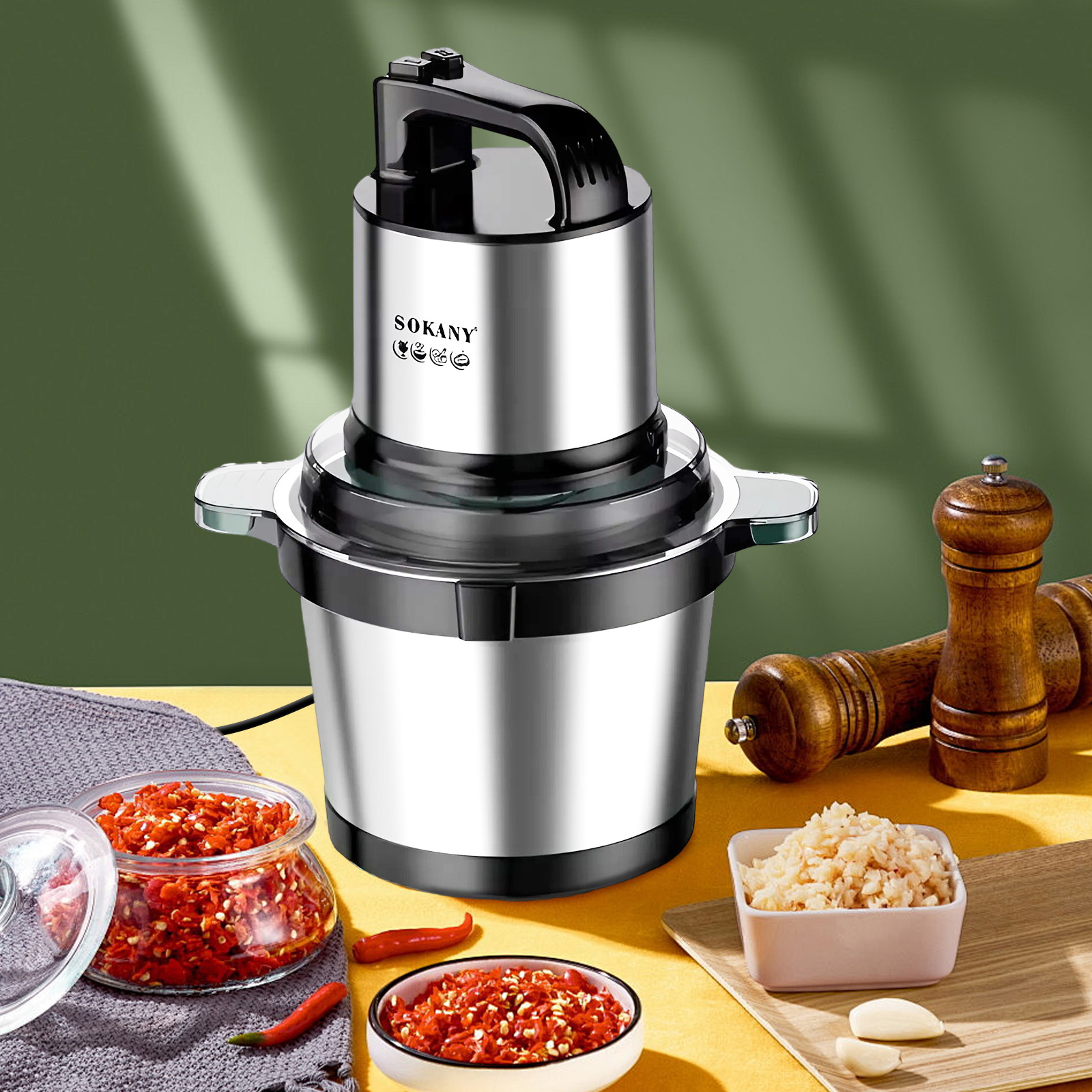 Robot da cucina argentato con manico nero su tavolo giallo. Si vedono ciotola di peperoni rossi e altri ingredienti.