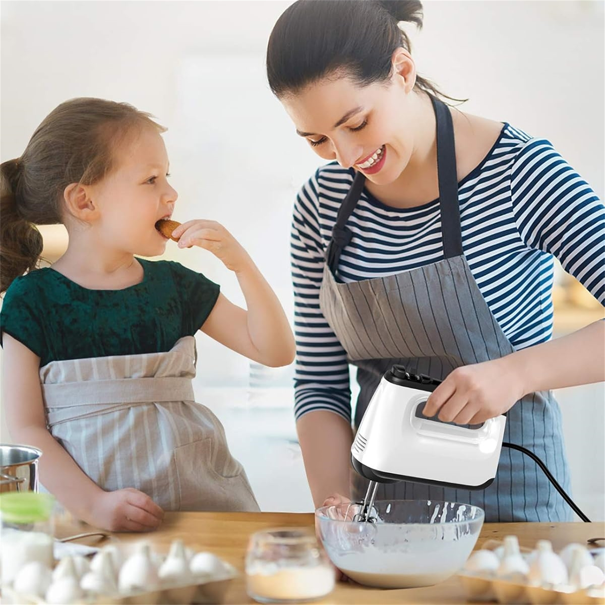 Frau mit weißem Handmixer, verrührt Zutaten in einer Schüssel. Ein Mädchen isst einen Snack in der Nähe.