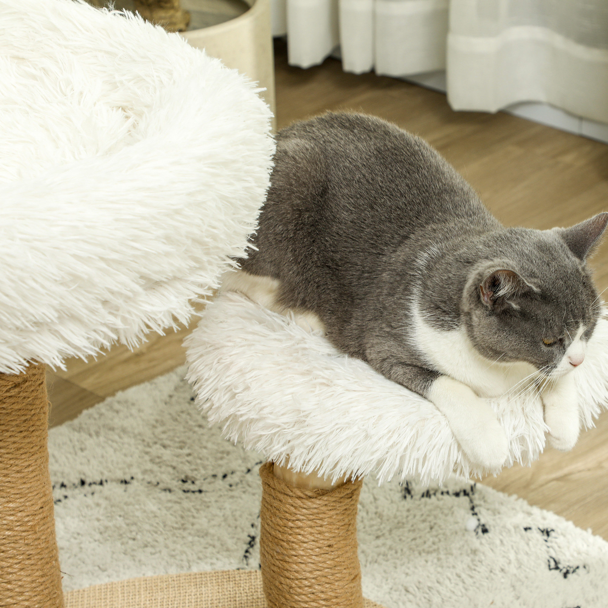 Eine grau-weiße Katze, die sich auf einem flauschigen, weißen Katzenbaum ausruht. Der Baum ist aus hellem Holz und Seil gefertigt.