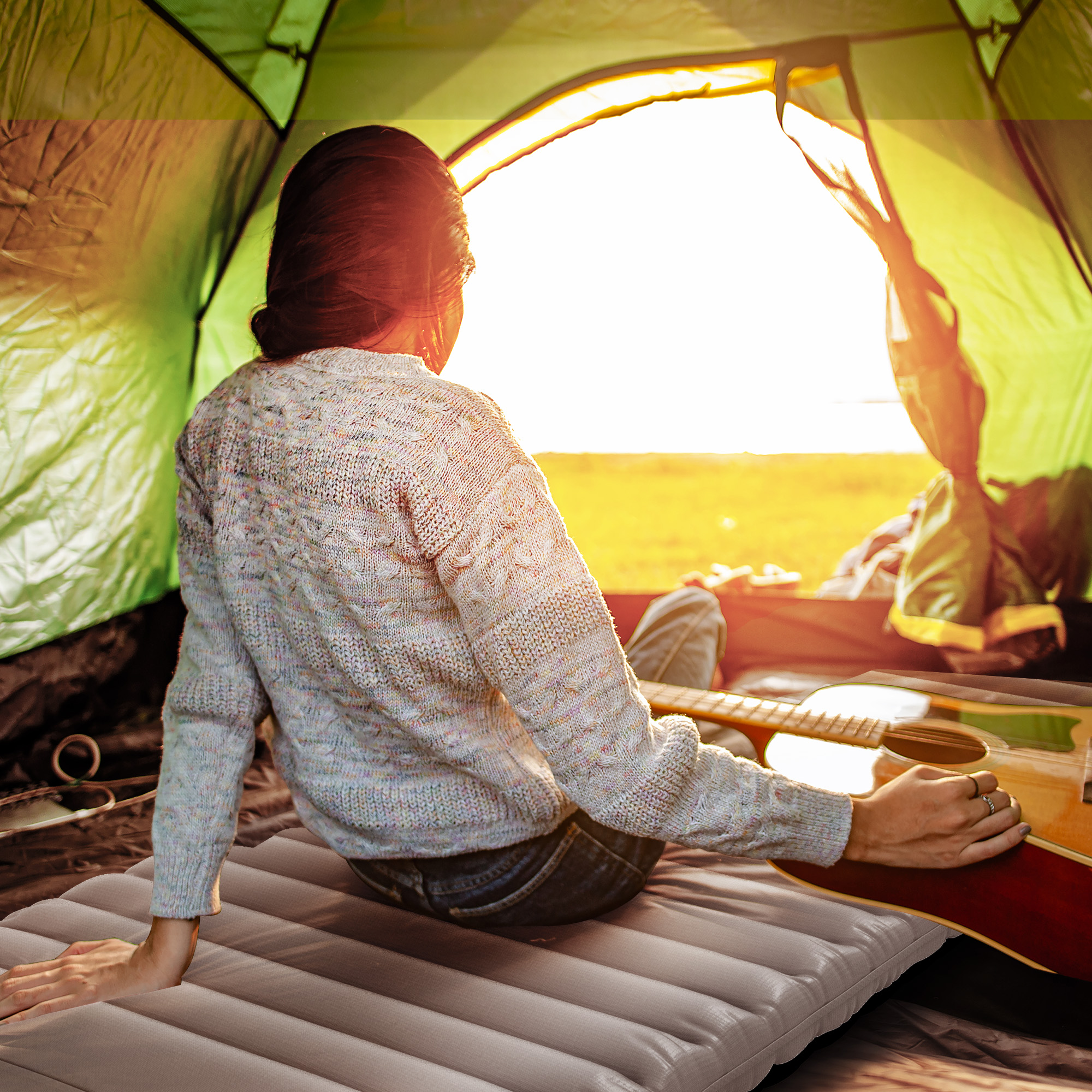 Frau sitzt in einem Zelt und blickt bei Sonnenuntergang auf ein Feld, mit einer Gitarre.