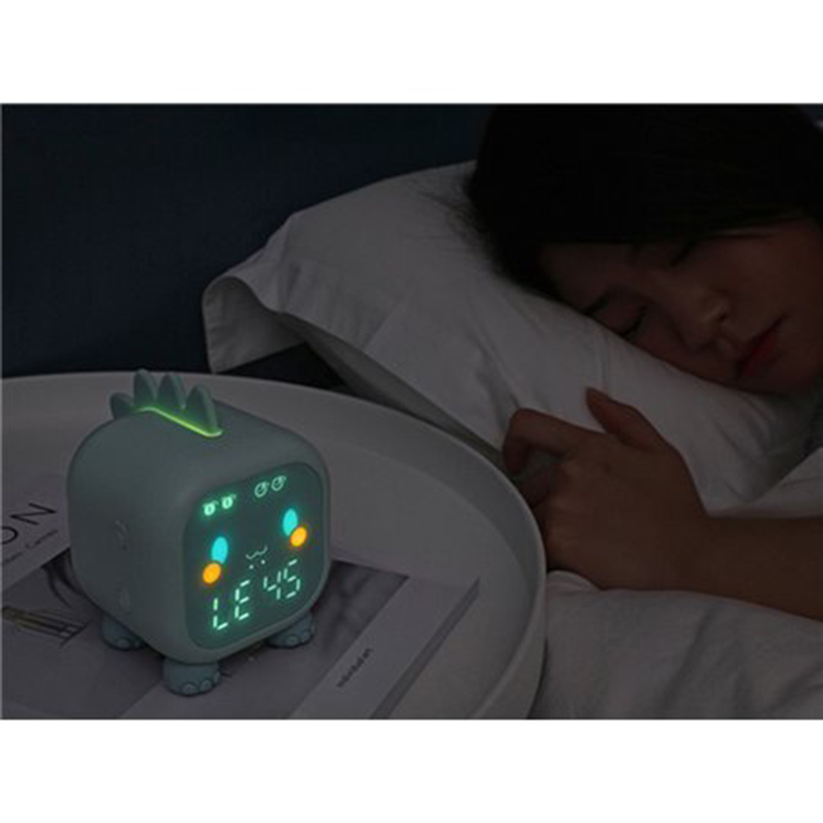Despertador con forma de dinosaurio en una mesita de noche. Una persona durmiendo al fondo.