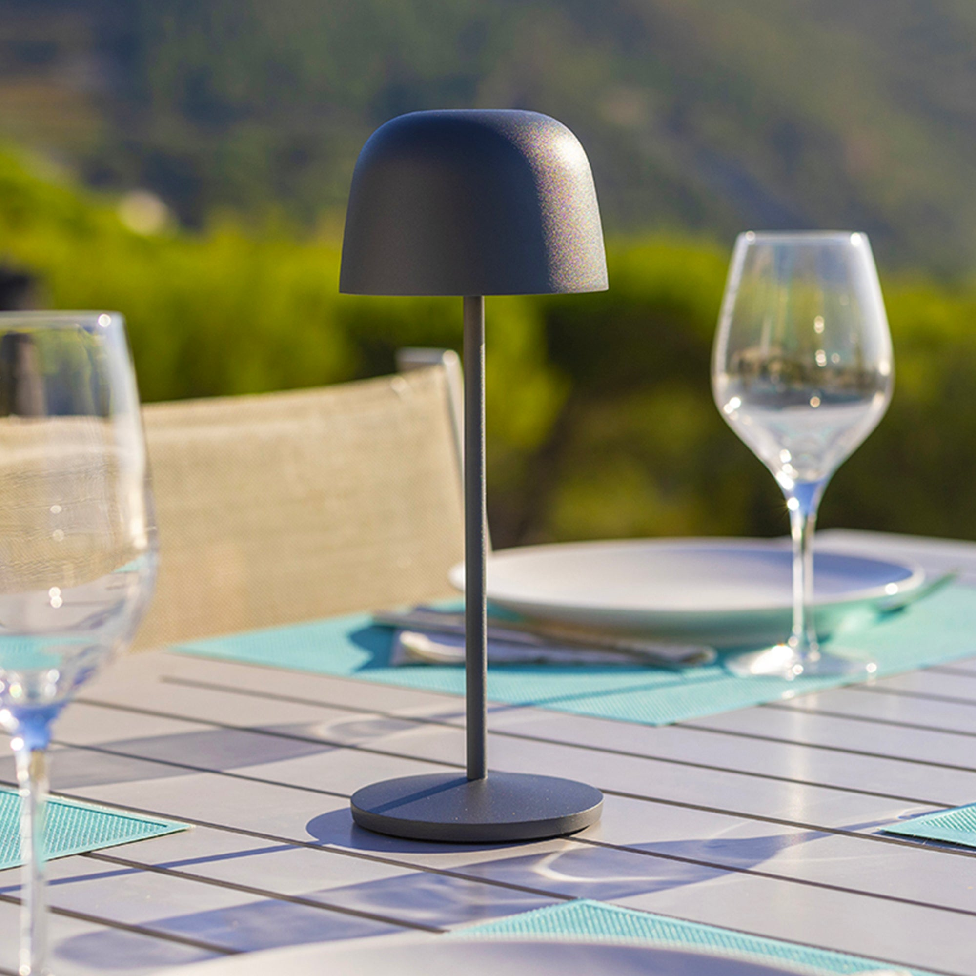 Eine dunkelgraue Tischlampe auf einem Tisch im Freien mit Gläsern und verschwommenem Hintergrund.