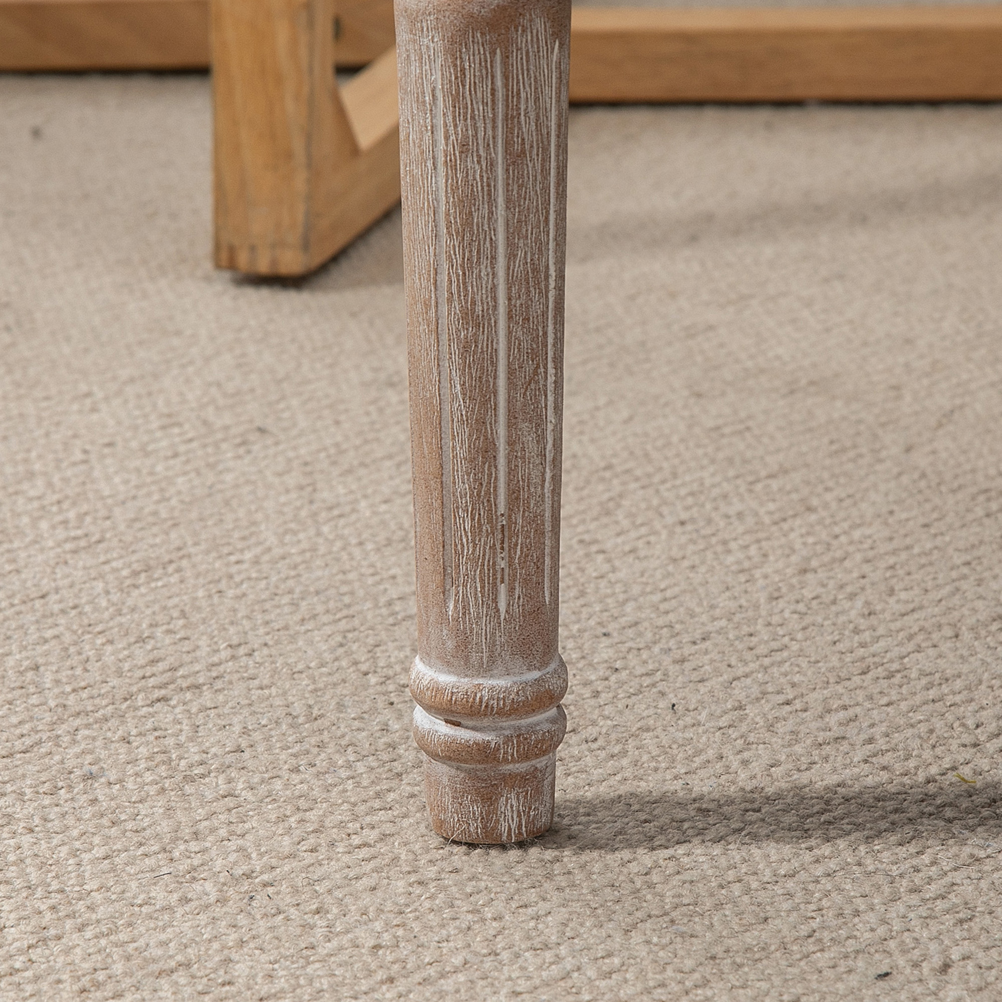 Nahaufnahme eines hellbraunen Holzbeins auf einem beigefarbenen Teppich. Ein Teil eines anderen Holztisches ist im Hintergrund.